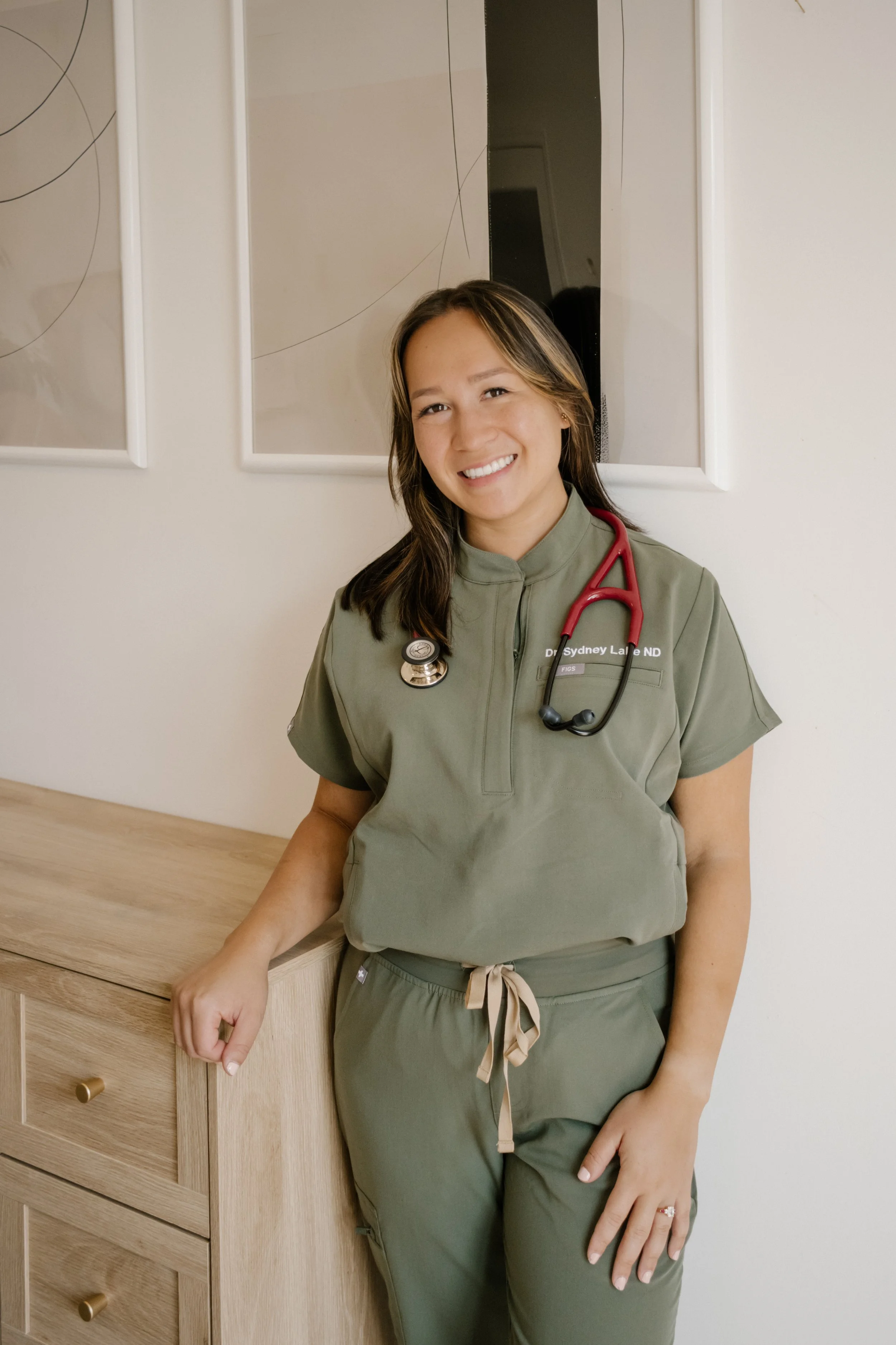 Dr. Sydney Lake, Naturopath in a Victoria, BC, in medical scrubs with a stethoscope around her neck, standing next to a wooden dresser and smiling at the camera.