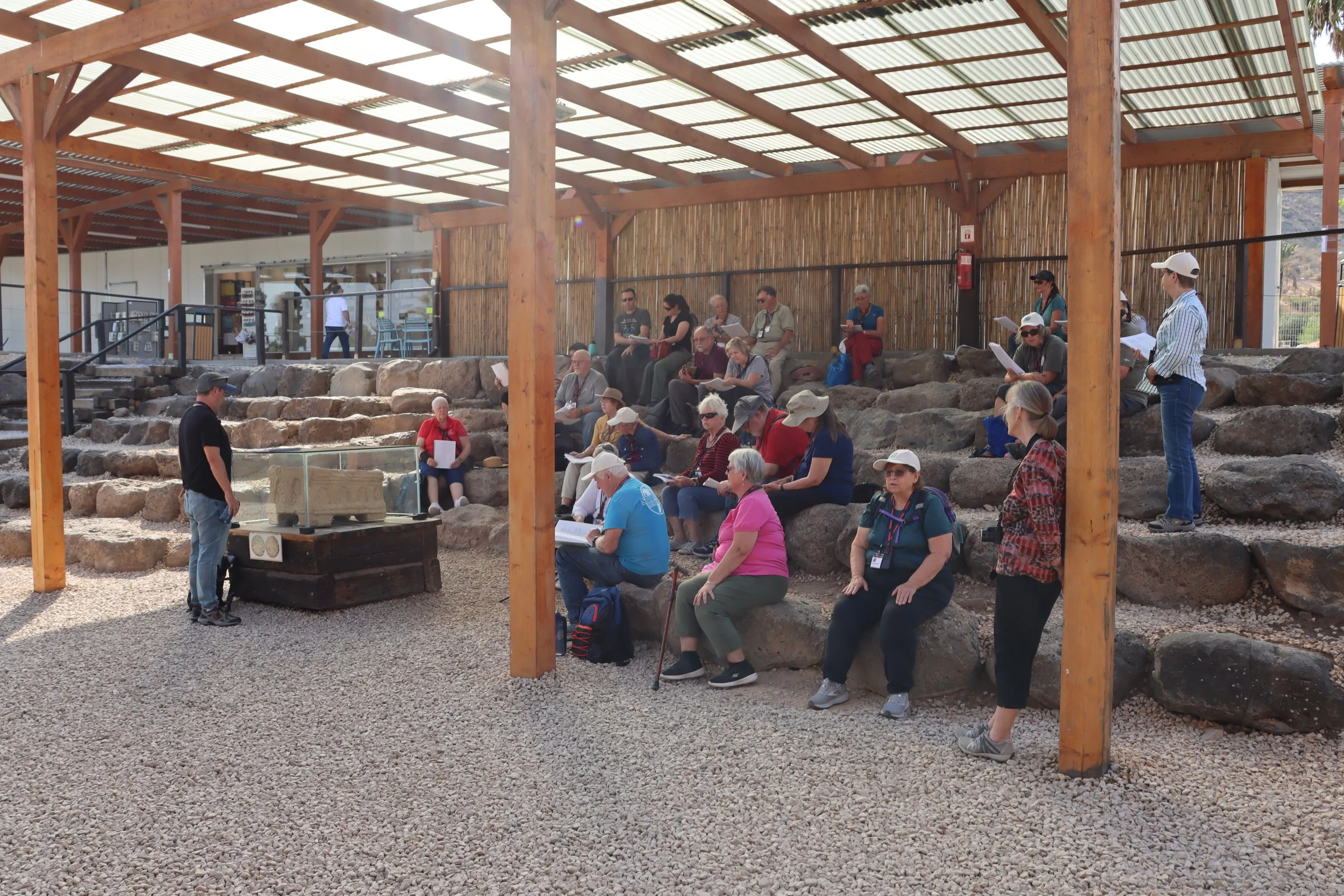 Group of people sitting and standing on stone tiered seating, some reading or looking at papers, under a wooden canopy with a bamboo fence backdrop, in an outdoor archaeological or historical site.
