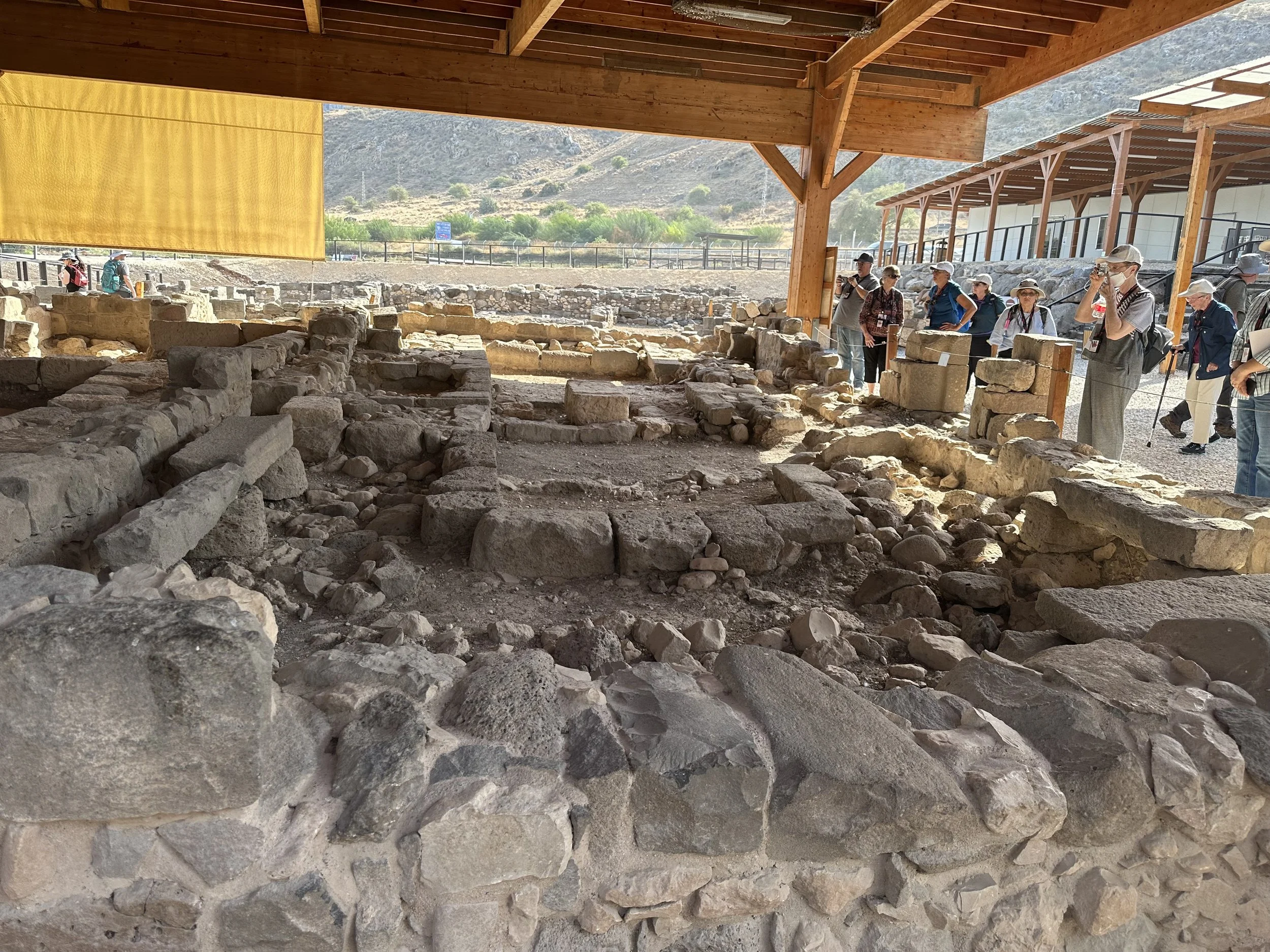 Archaeological excavation site with ancient stone ruins under a wooden roof, and a group of tourists observing and taking pictures.