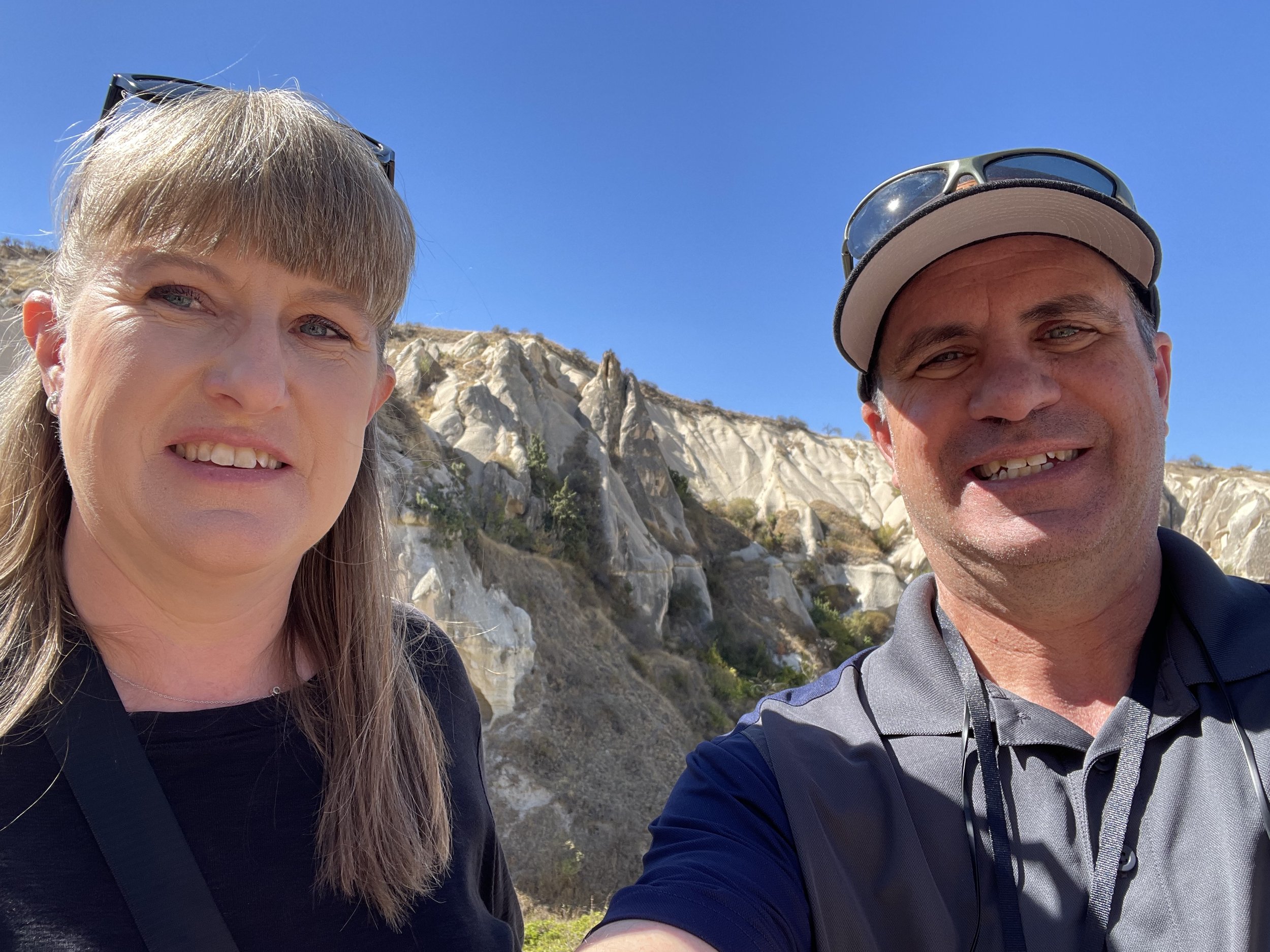 A smiling middle-aged woman with blonde hair and a man with sunglasses on his cap, both outdoors against a rocky hillside and clear blue sky.
