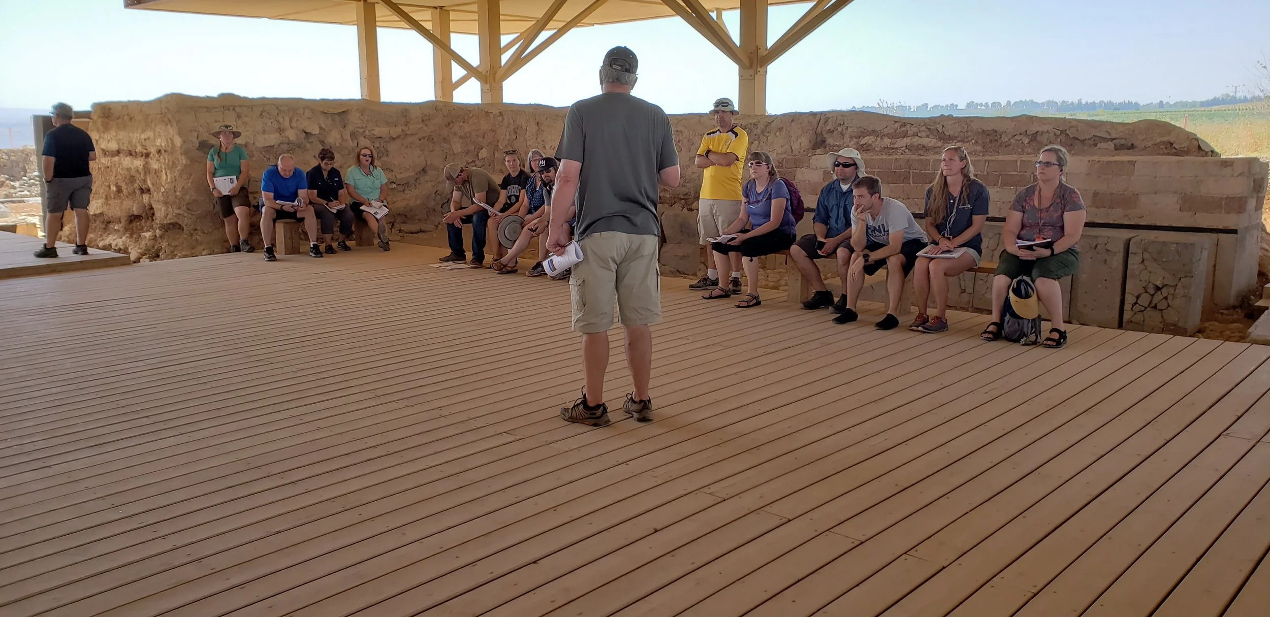 Group of people sitting on benches and listening to a tour guide at an archaeological site with ancient brick and mud walls, under a wooden shelter.