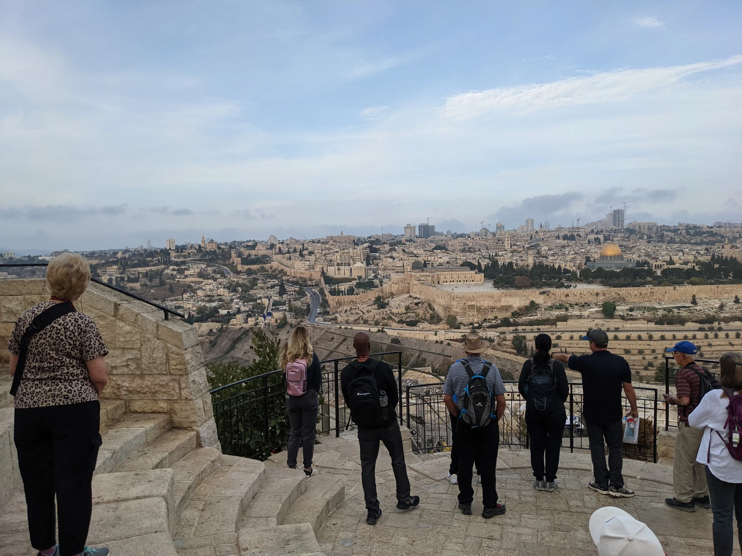Tourists viewing a panoramic cityscape of Jerusalem from a historic viewpoint, with some taking pictures and others looking out over the city.