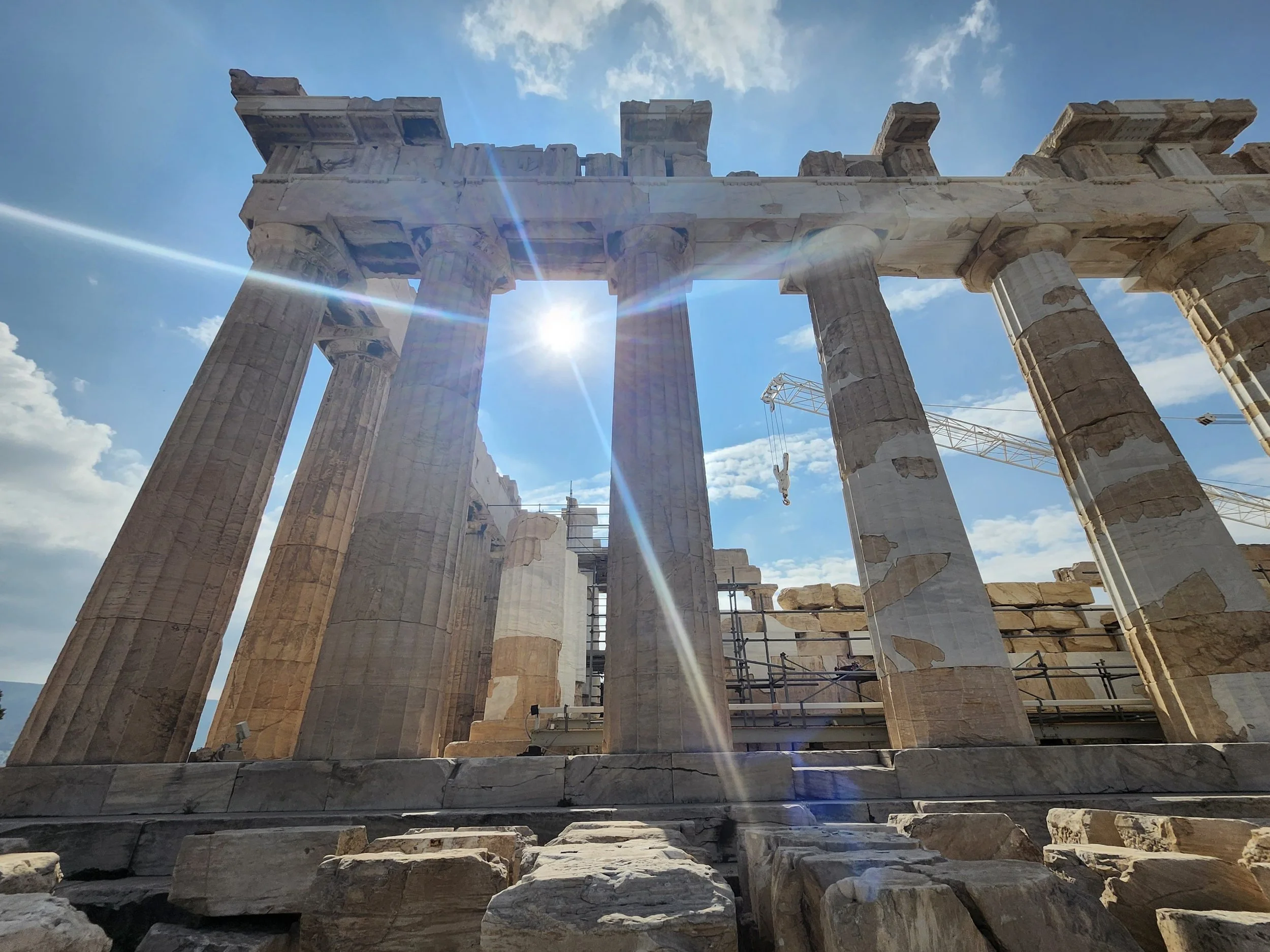 Ancient Greek ruins with tall stone columns against a bright blue sky with clouds and sunlight.