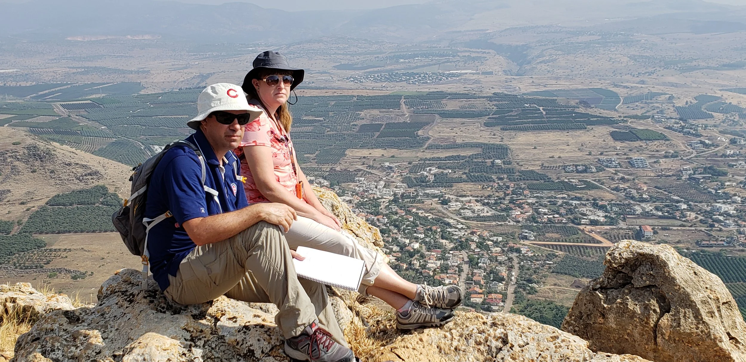 Two people sitting on rocks at a mountain summit overlooking a valley with hills, farmland, and a small town.