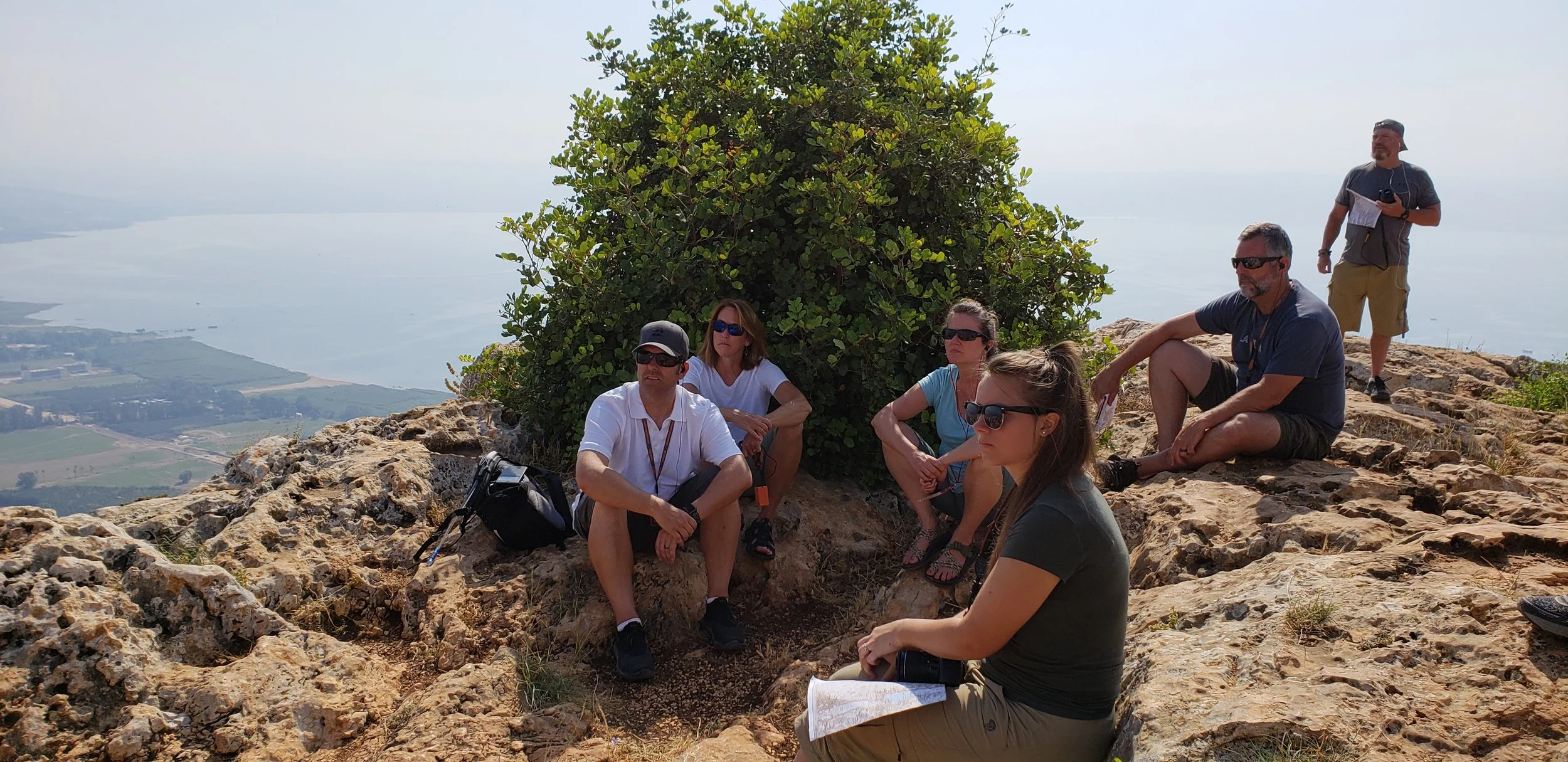 Group of people resting on rocky terrain at a high vantage point with a large bush behind them, overlooking a body of water and landscape below.