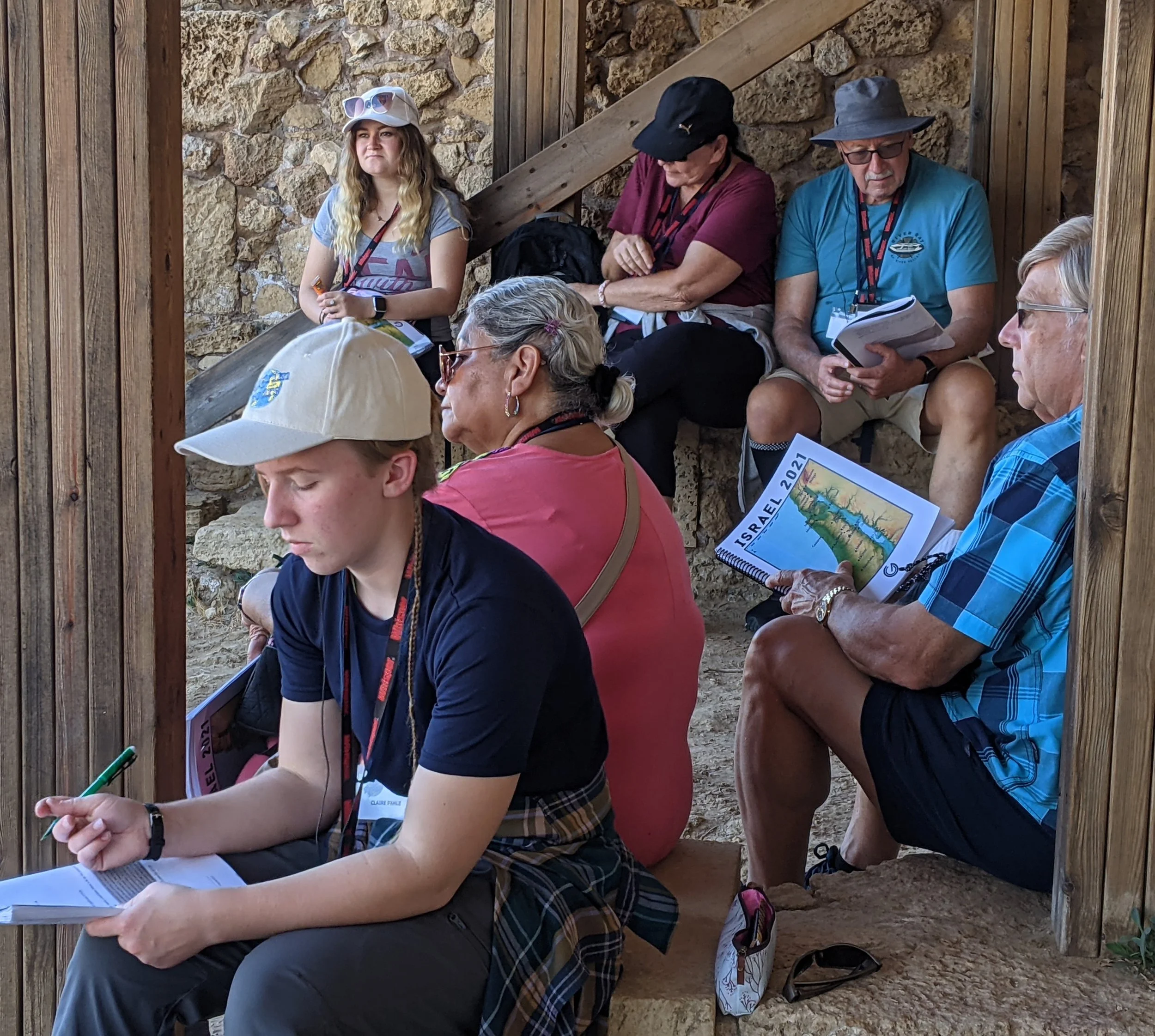 Group of people sitting under wooden and stone structure, some reading or writing, others engaged in conversation, with outdoors setting.