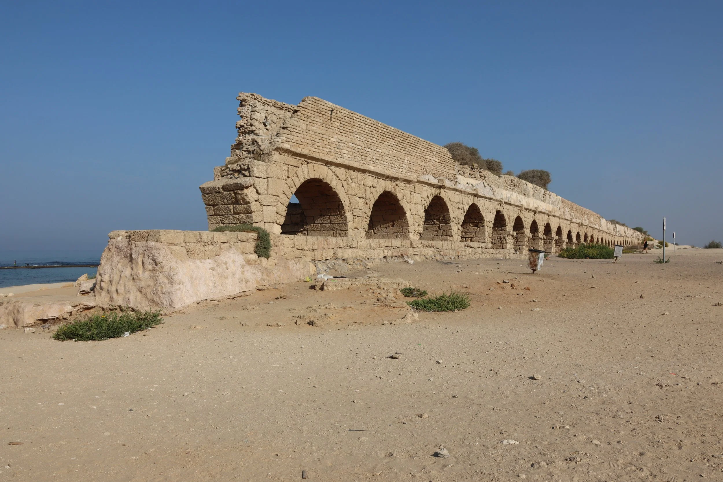 Ancient stone aqueduct with multiple arches extending along a sandy landscape under a clear blue sky.