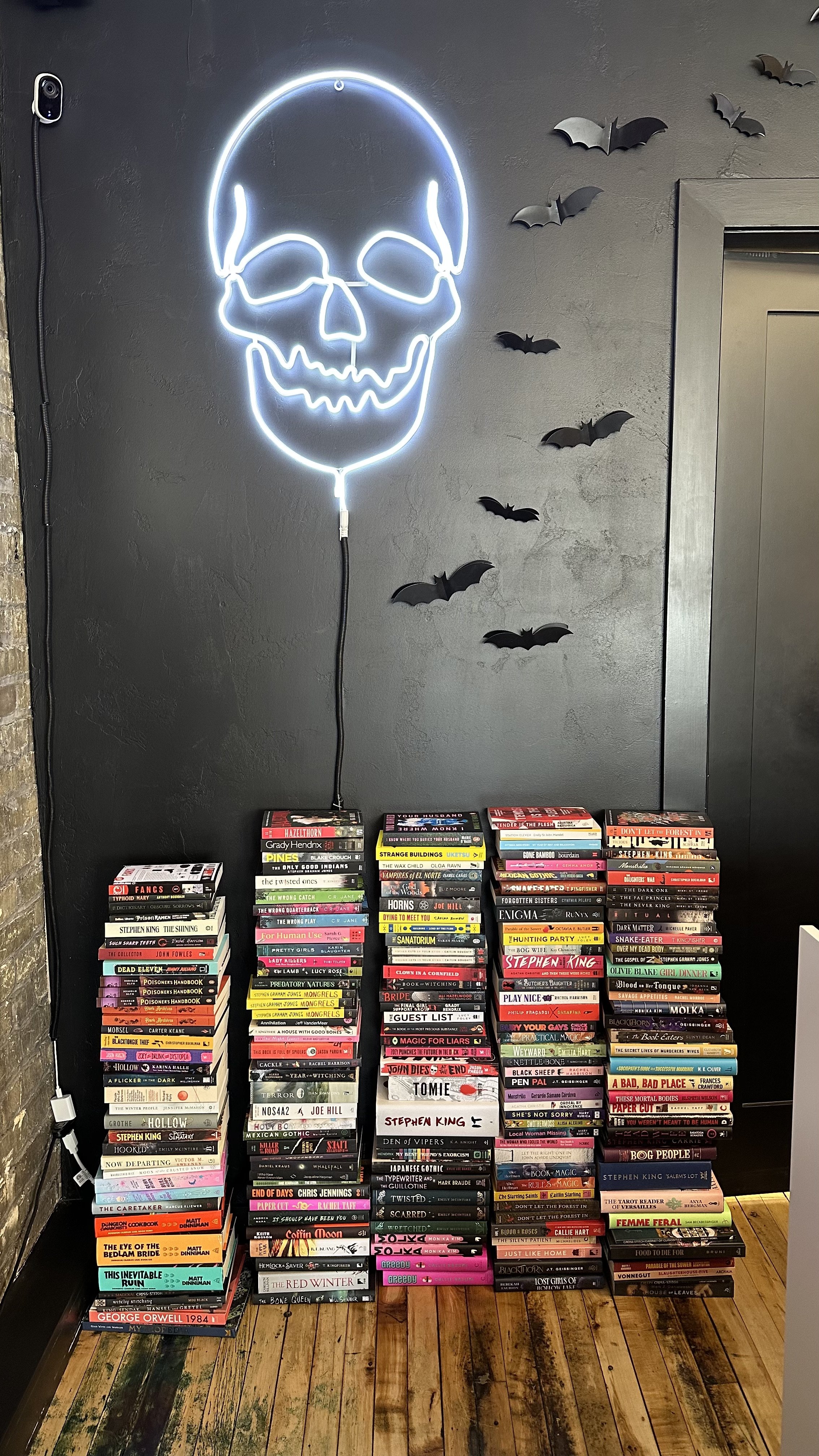 A black wall decorated with a white neon skull and black paper bats. Below are stacks of paperback books organized on the floor.