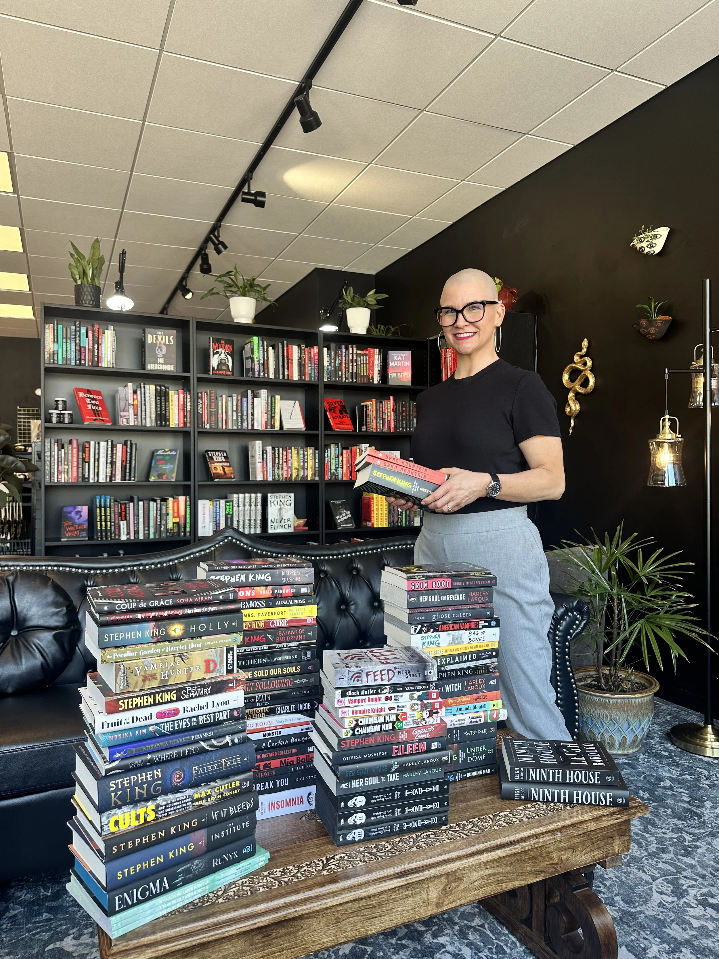 A woman with glasses and a shaved head is standing in a bookstore surrounded by stacks of books, holding three books in her hands. The background features black bookshelves filled with books and plants on top, along with decorative wall art. The setting is modern with a black and dark gray color scheme.