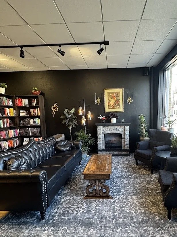 A cozy living room with black leather sofas, a wooden coffee table, a dark accent wall with a fireplace, bookshelves filled with books, and various plants. A large window on the right lets in natural light.