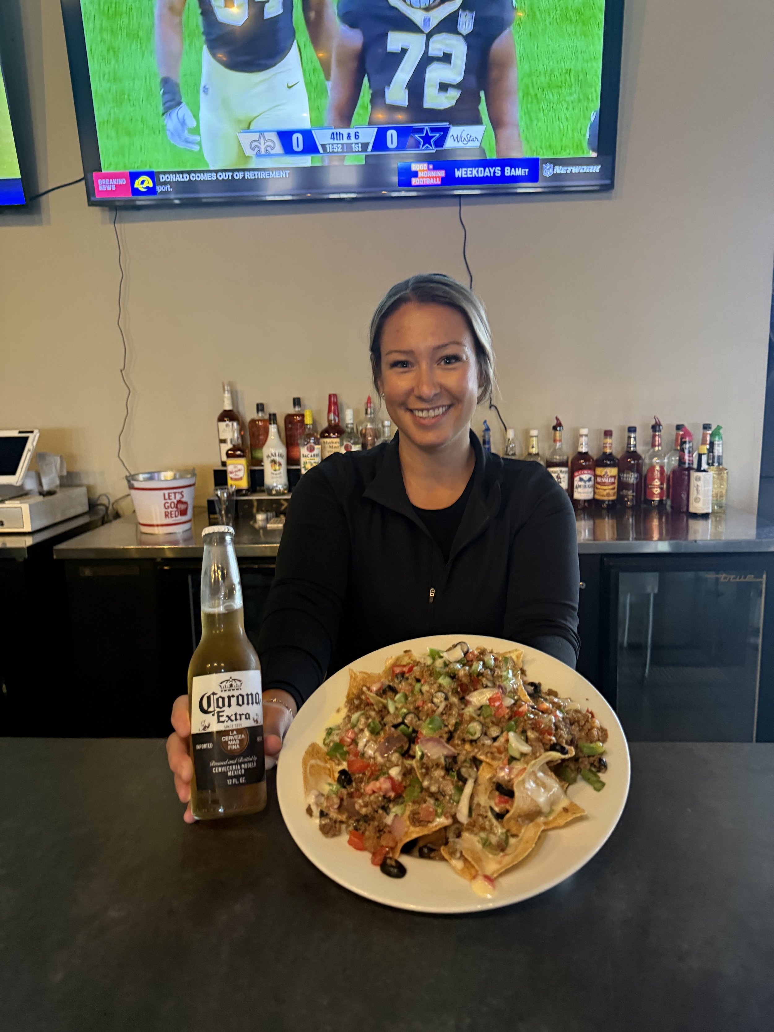 A woman smiling at camera holding a plate of nachos topped with vegetables and cheese, and a bottle of Corona Extra beer in front of her. She is seated at a restaurant with a bar and liquor bottles behind her, and a TV displaying a football game in the background.