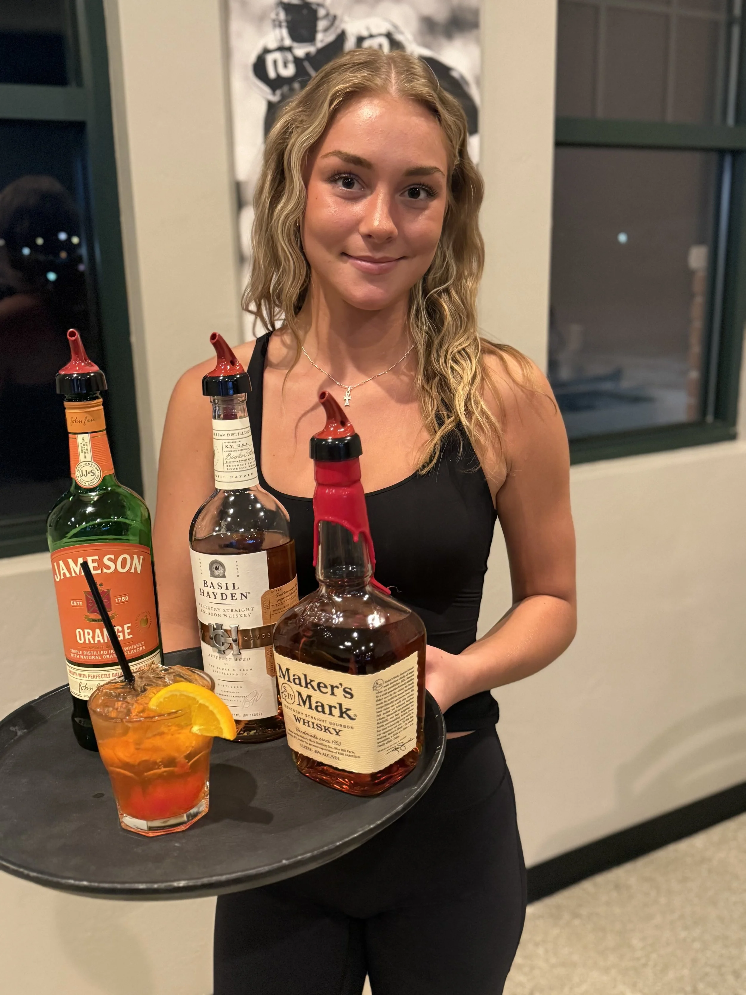 A woman with wavy blonde hair and a black sleeveless top holding a black tray with bottles of alcohol, a glass of orange cocktail with an orange slice, in a room with a large window and a black and white poster on the wall behind her.