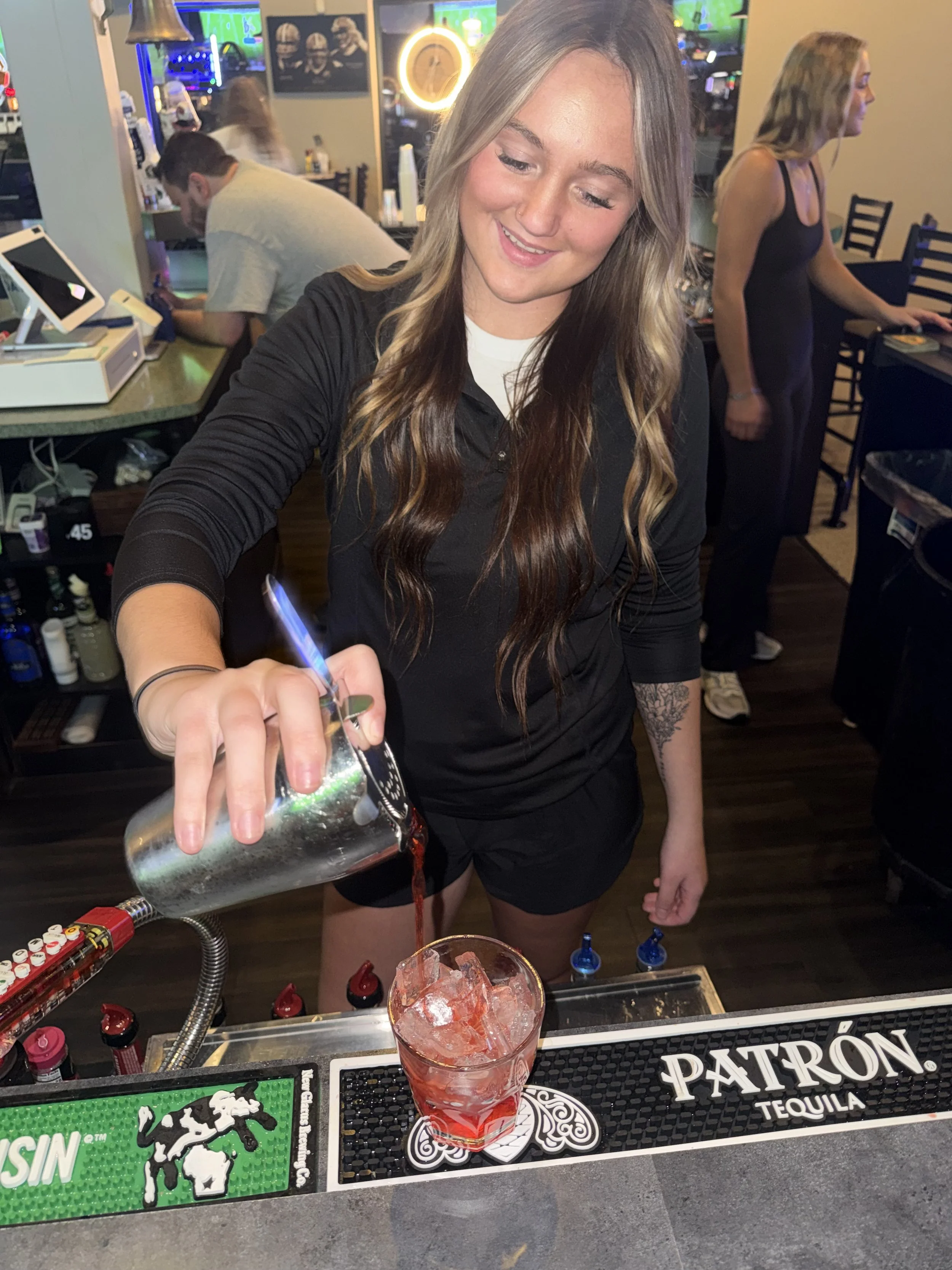A woman with long, wavy hair wearing a black top and shorts is pouring a drink from a shaker into a glass with ice at a bar. In the background, a man is sitting at the bar counter, and a woman in a black dress is working behind the bar. There are bar