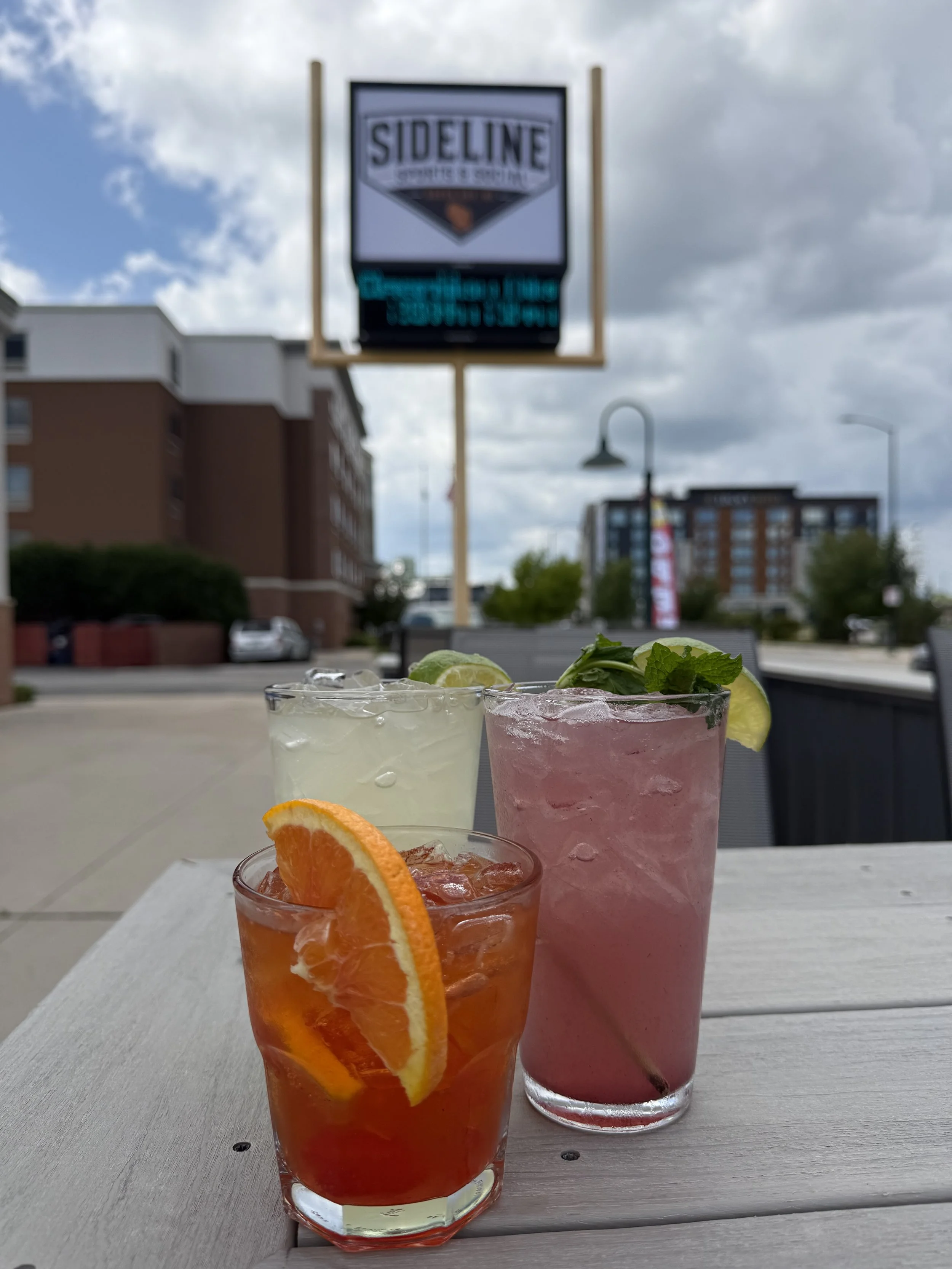 Three colorful cocktails on a white table outside with a cloudy sky and a billboard in the background. The drinks are garnished with slices of orange, lemon, and mint leaves.