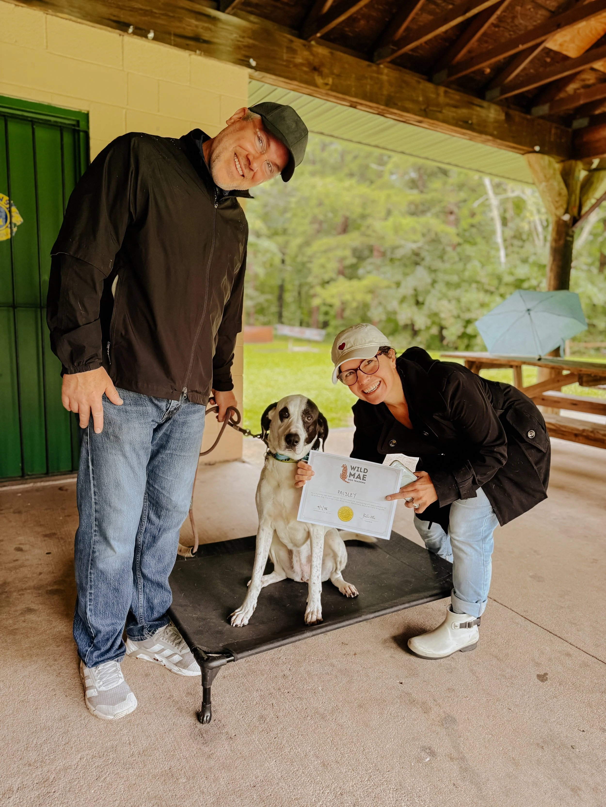 A man and a woman are smiling and posing with a dog that has a certificate, in an outdoor setting with trees in the background. The woman is holding the certificate, and they are near a green door, under a wooden and metal roof.