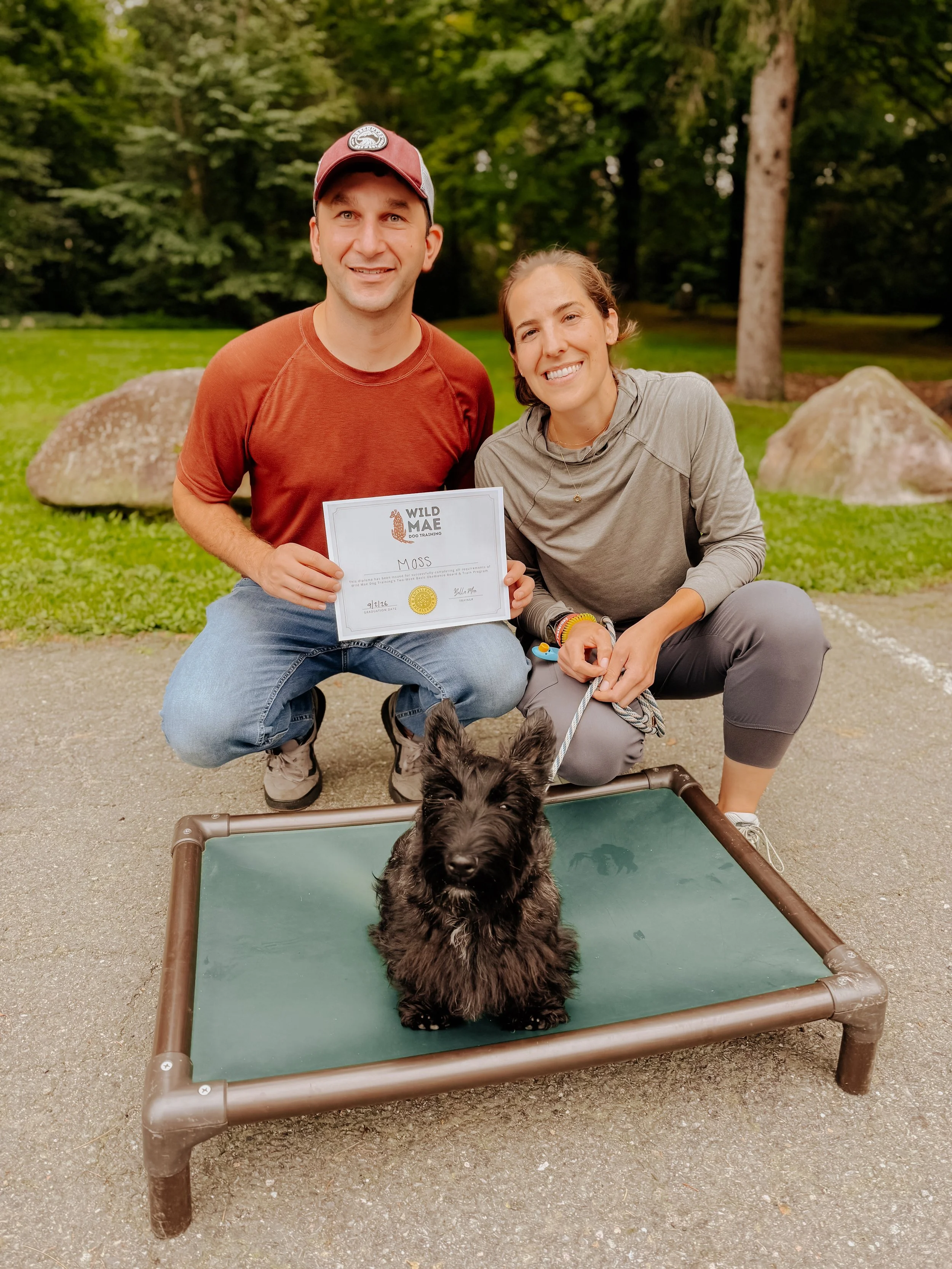 A man and woman kneeling next to a black dog on a leash, with a certificate in the man's hand. They are outdoors in a park with trees and grass, and a small dog bed in the foreground.