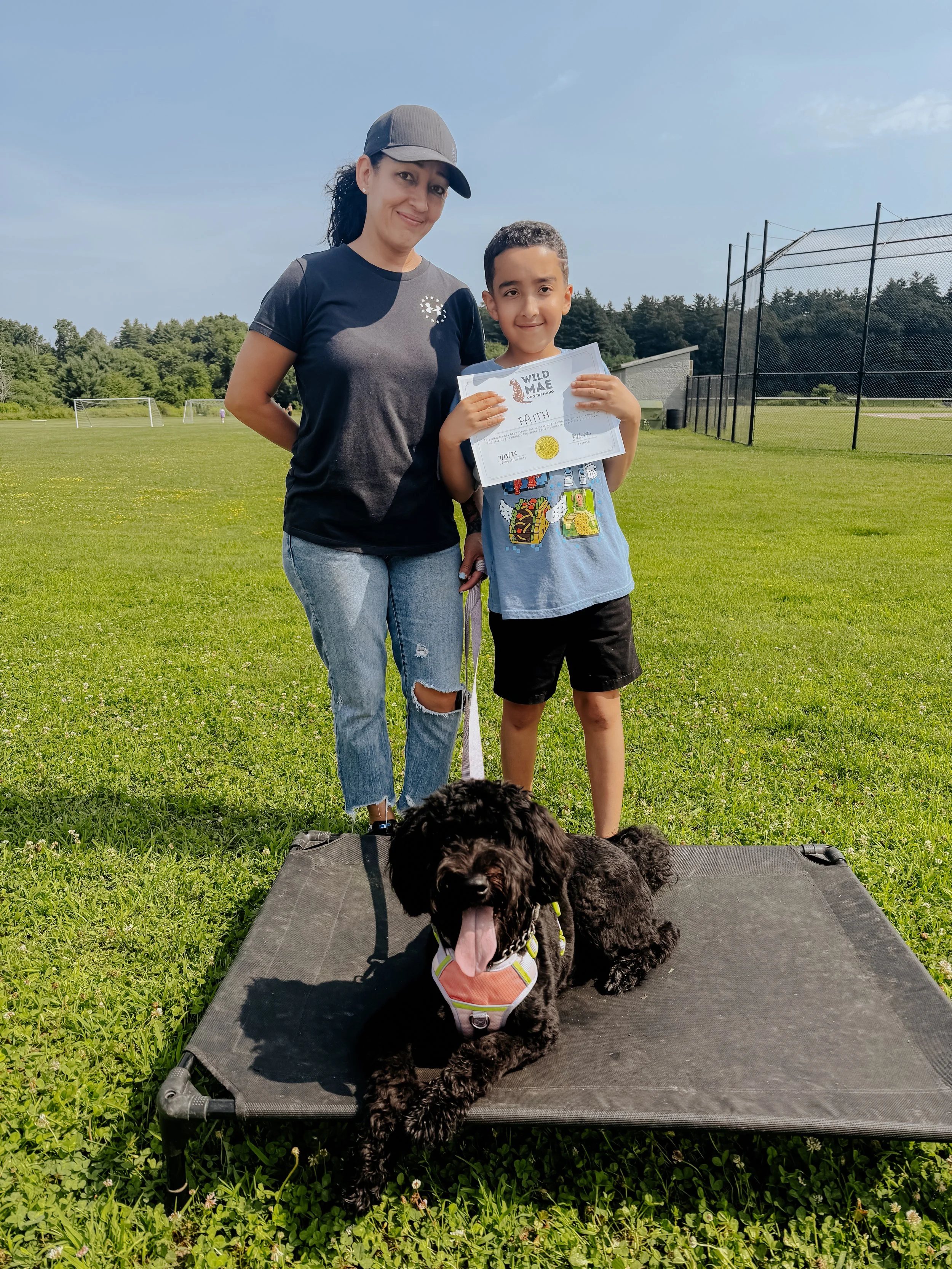A woman and a young boy standing in a grassy field with soccer goals in the background. The boy is holding a certificate that says 'Faith' and has a gold seal. There is a black dog lying on a raised platform in the foreground, wearing a vest and pant