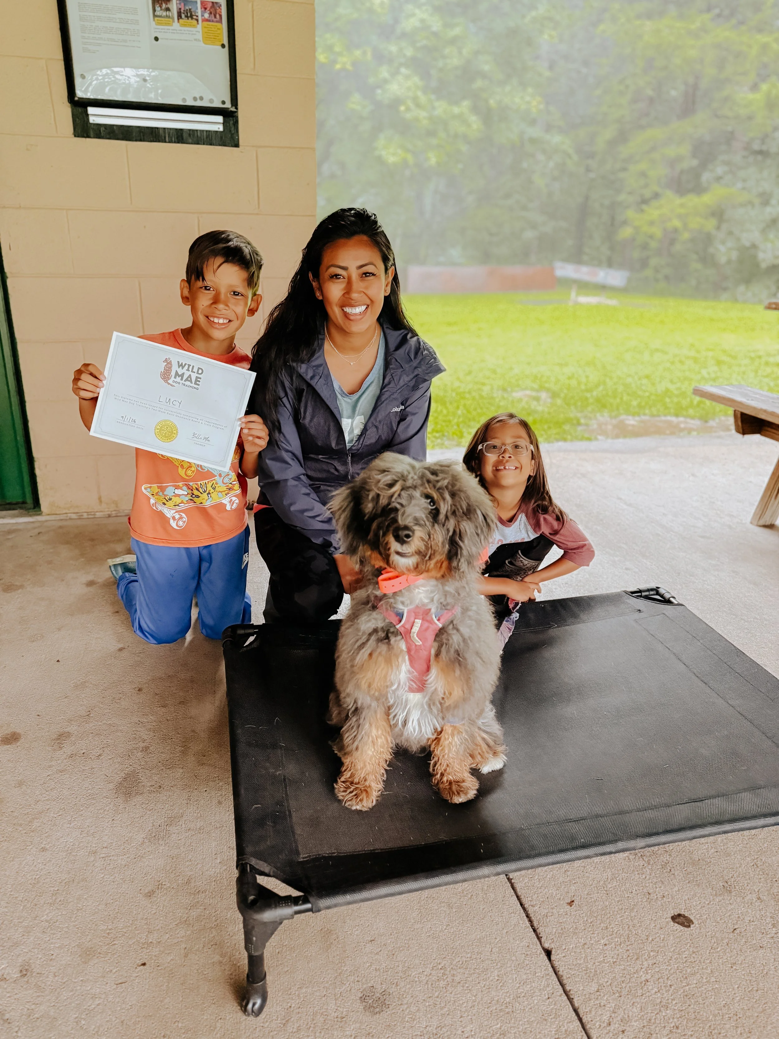 A group of three children and a dog in a park or outdoor shelter. One boy is holding a certificate, and two girls are sitting or kneeling on the ground. The dog is a fluffy, large breed with a pink harness, sitting on a black platform. The background