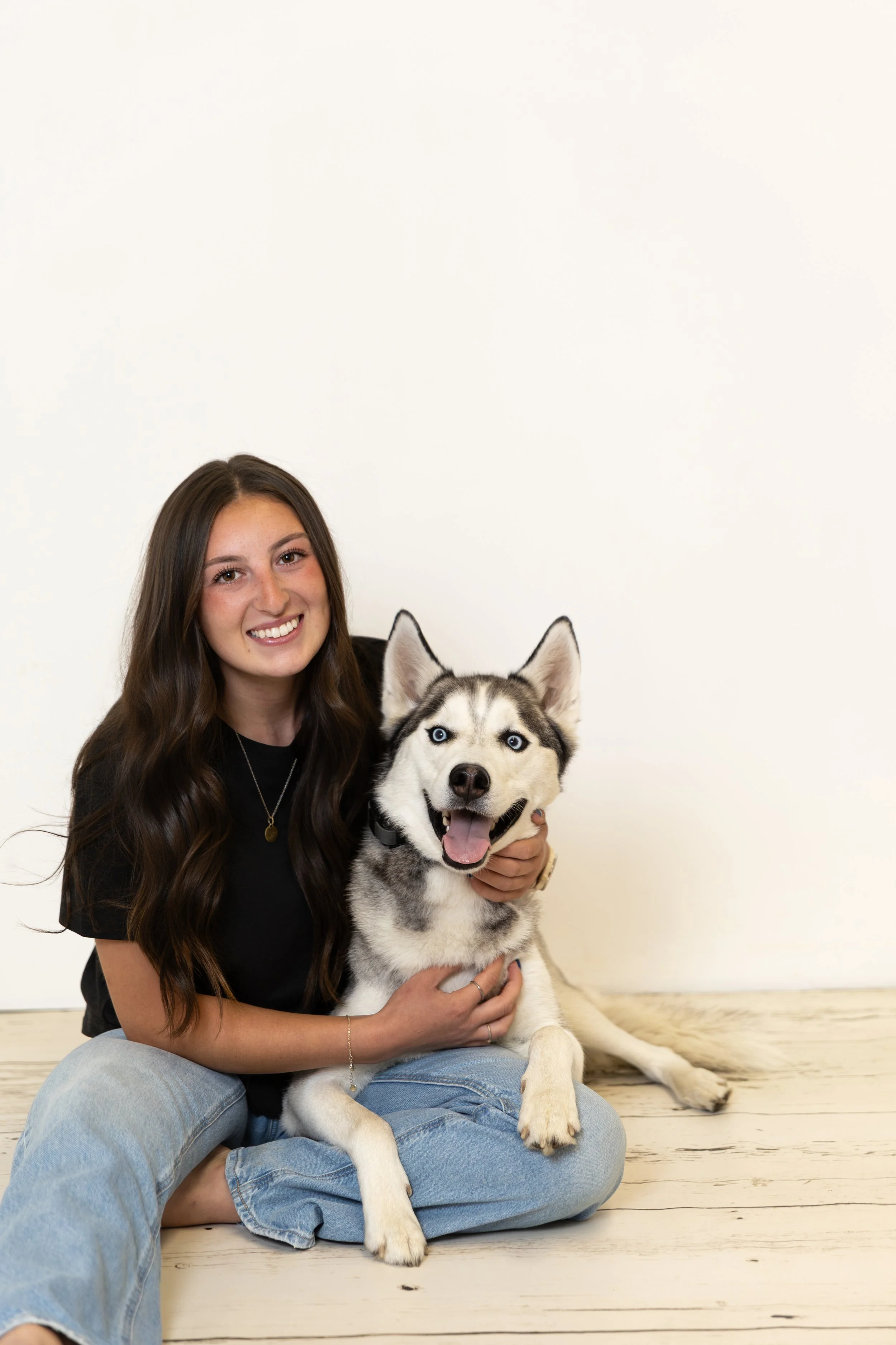 A young woman with long, wavy brown hair smiling while holding a happy Siberian Husky puppy with blue eyes. They are sitting on a wooden floor against a plain white background.