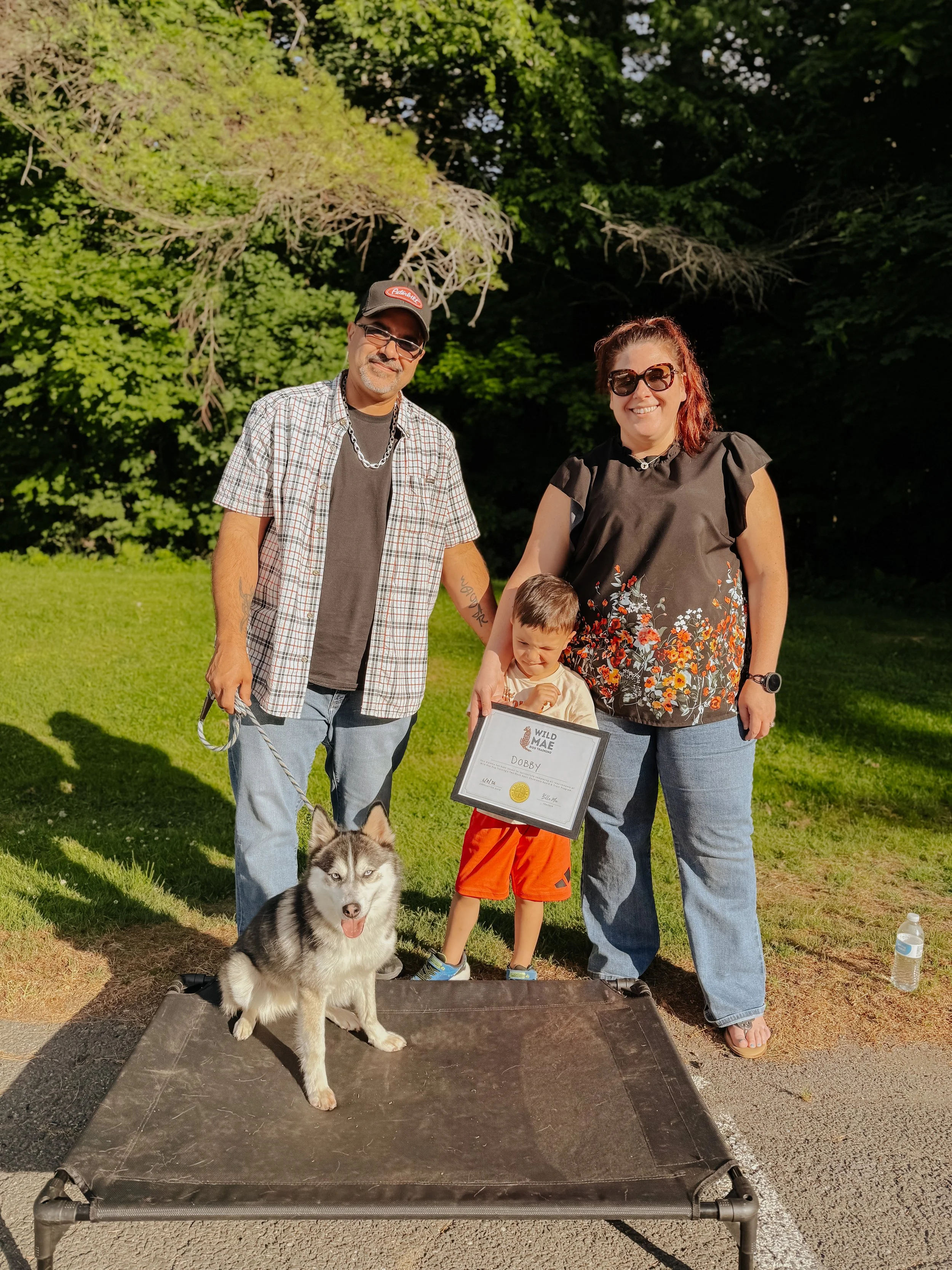 A family with a Siberian Husky dog on a winner's podium at an outdoor event. The boy holds a framed certificate, and the dog is sitting on the ground in front of the podium. The parents stand behind the boy, smiling, with a background of green trees 