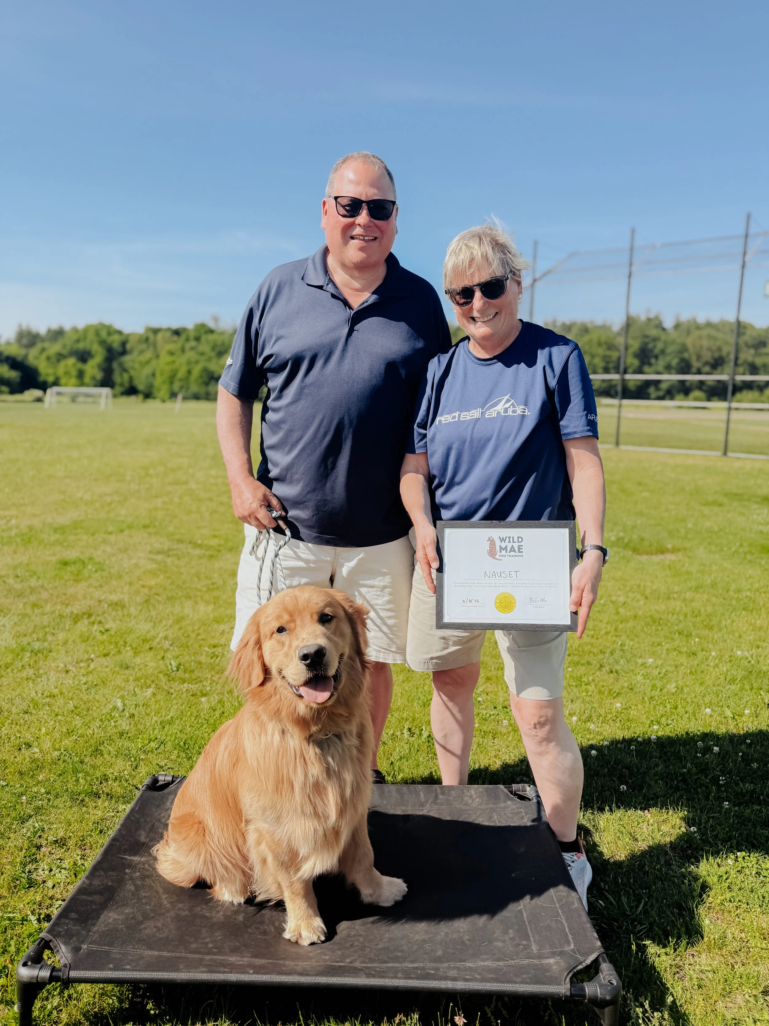 A man and woman stand outdoors on a grassy field, smiling, holding a framed certificate that reads 'NAUSET', with a golden retriever puppy sitting in front of them. The man is wearing a dark polo shirt and sunglasses, and the woman is wearing a navy 