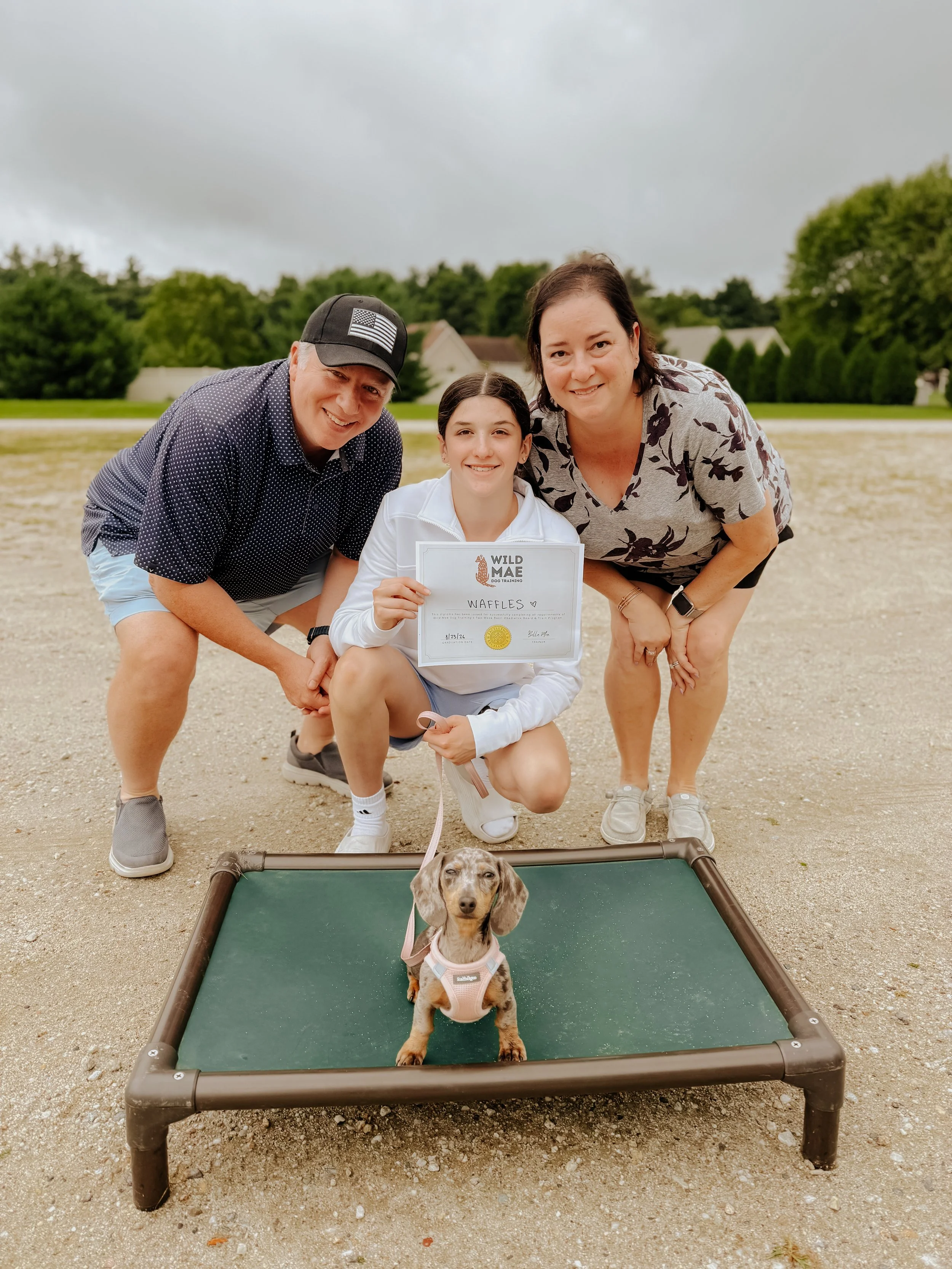 Three people kneeling outdoors behind a small dog on a green platform, holding a certificate. The dog is a dachshund wearing a pink harness, and there is a cloudy sky and trees in the background.