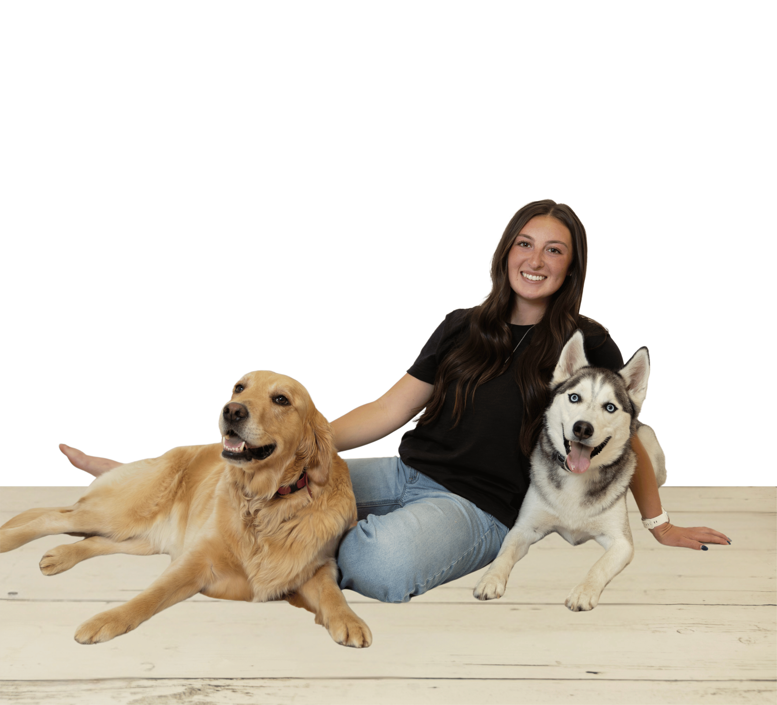 A young woman with long brown hair sitting on a wooden floor with two dogs, a Siberian Husky and a golden retriever, both lying beside her.
