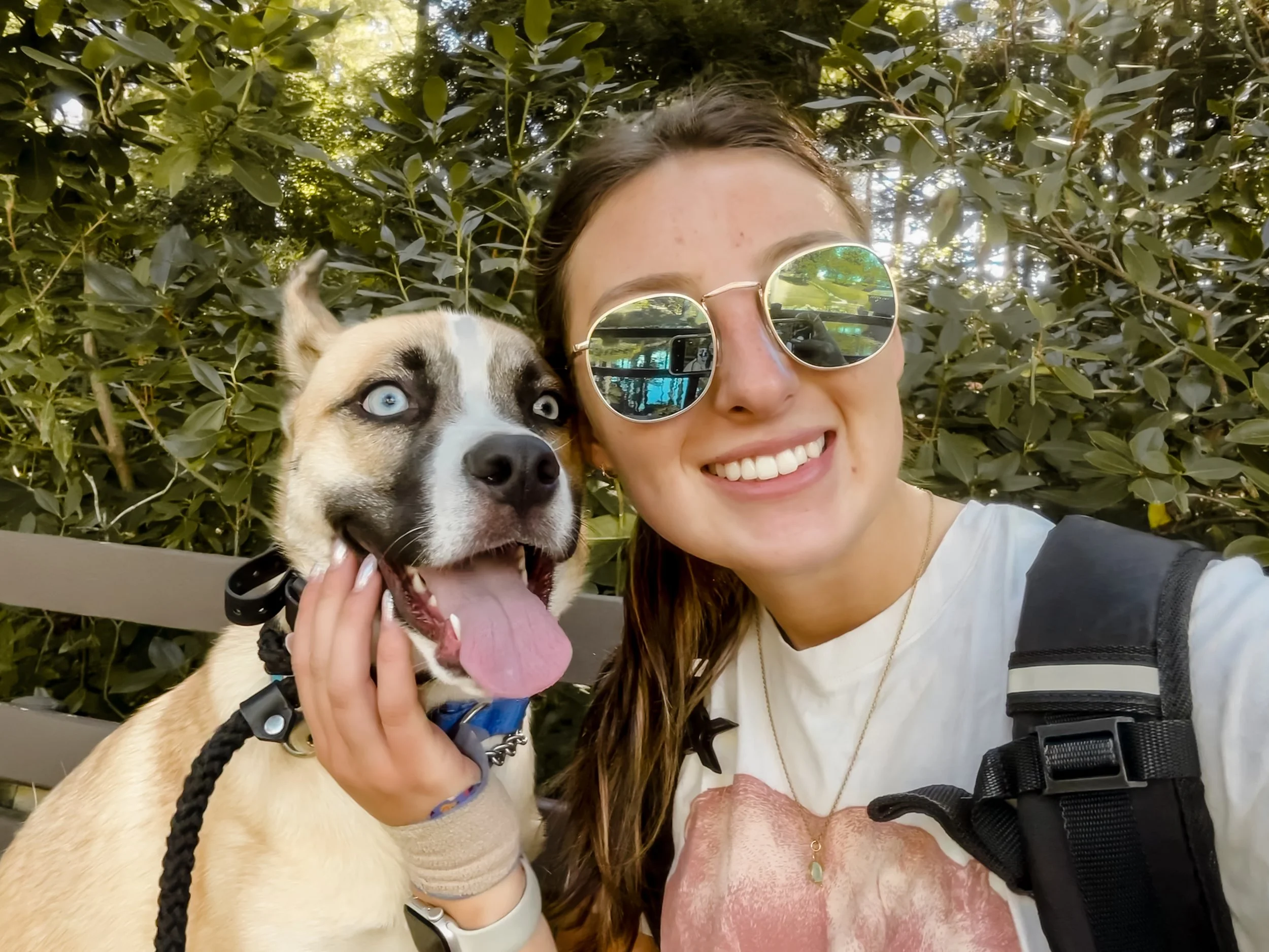 A young woman with long reddish-brown hair and reflective round sunglasses taking a selfie with a Siberian Husky dog outdoors in front of green leafy bushes.