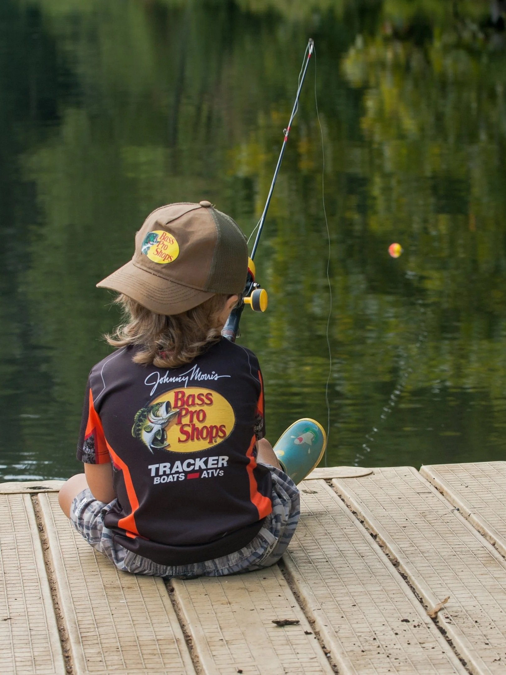Young boy sitting on a dock fishing on a calm lake.