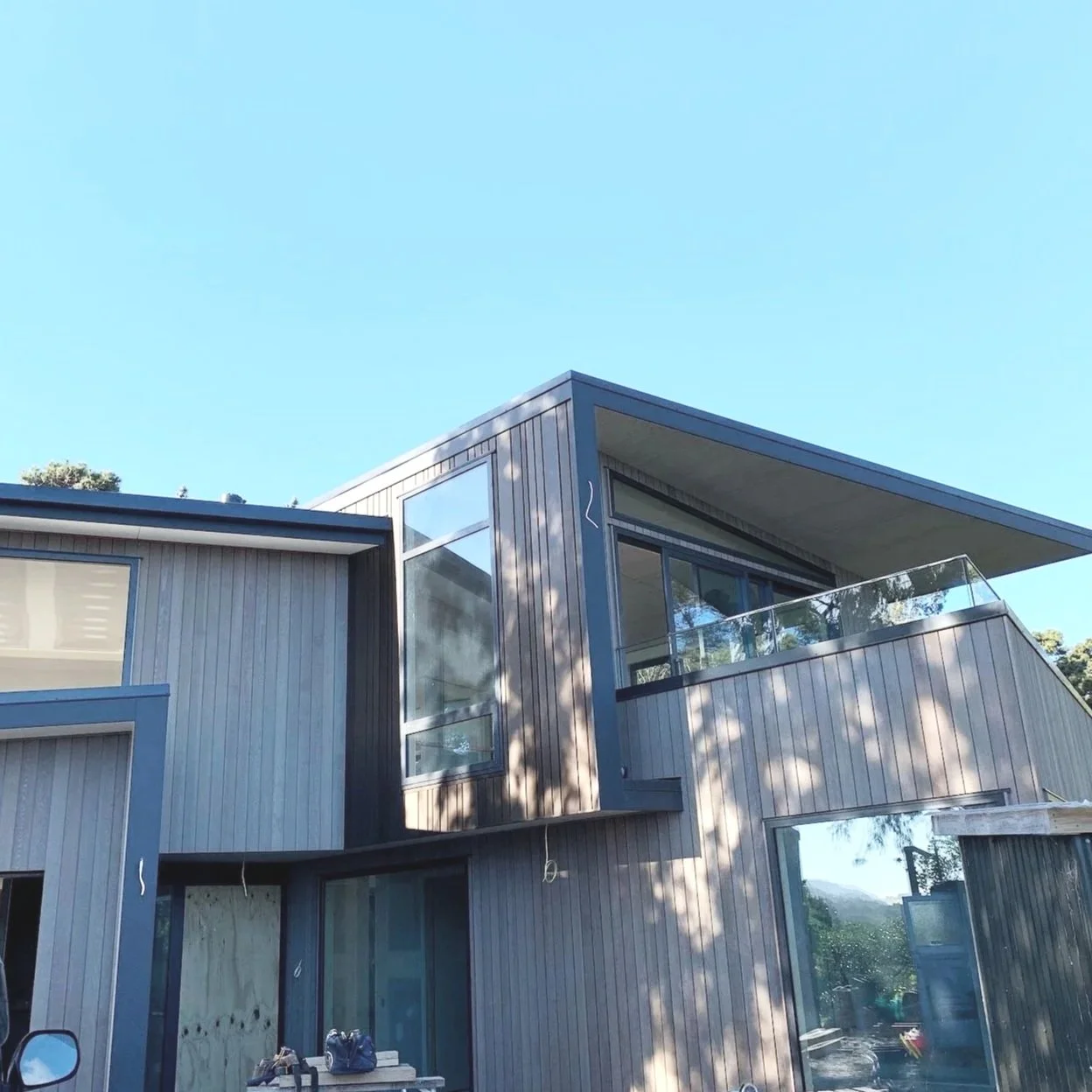 Modern multi-story house with large windows, wood siding, and a glass balcony, under a clear blue sky.