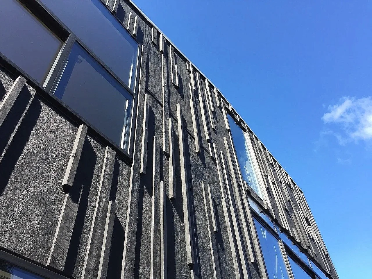 Looking up at a modern building with an irregular facade of vertical black panels and large reflective windows under a clear blue sky.