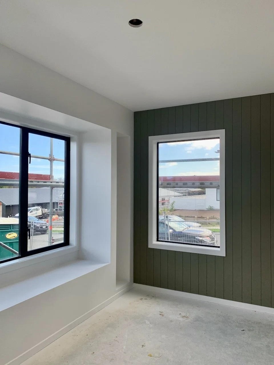 Interior view of a room under construction with two large had painted windows, white and dark green walls, and an unfinished concrete floor.