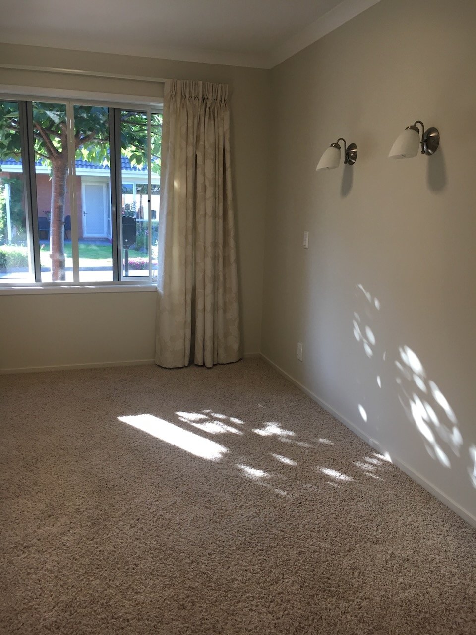 Empty room with beige carpet, off-white walls, a window with curtains, and sunlight creating patterned shadows on the floor and wall.