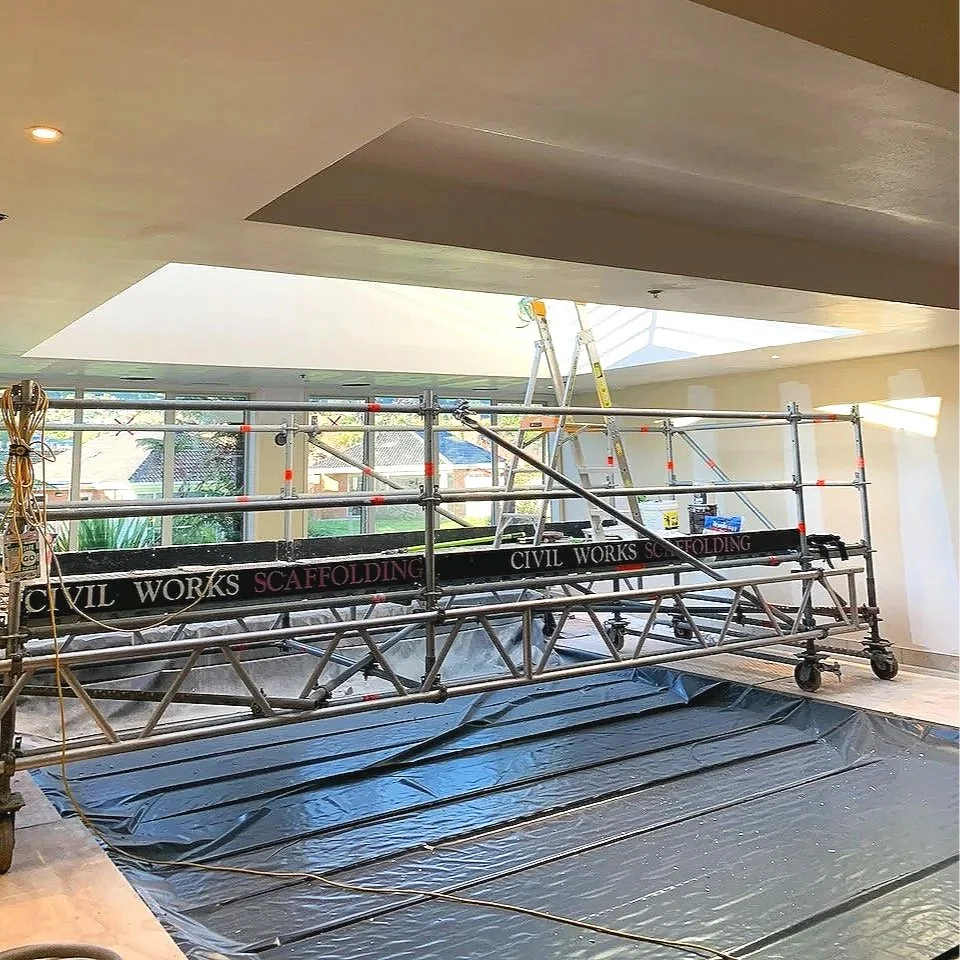 Indoor construction site with scaffolding labeled 'Civil Works Scaffolding' and a ladder, plastic sheeting on the floor, and a large opening in the ceiling allowing natural light.