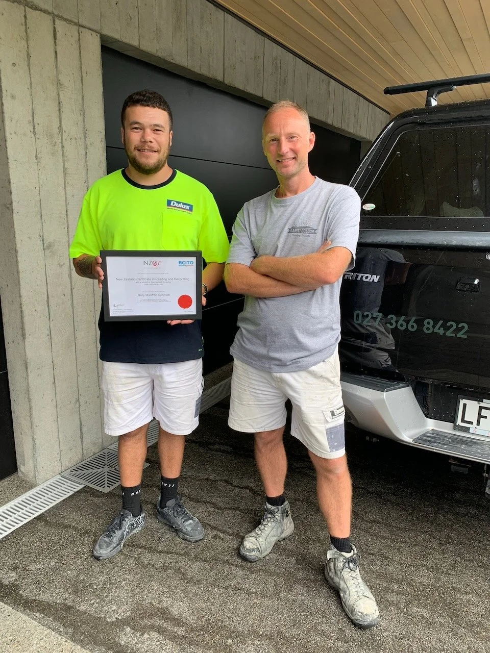 Two men standing outdoors, one holding a certificate, both dressed in casual work clothes, smiling, next to a black vehicle.