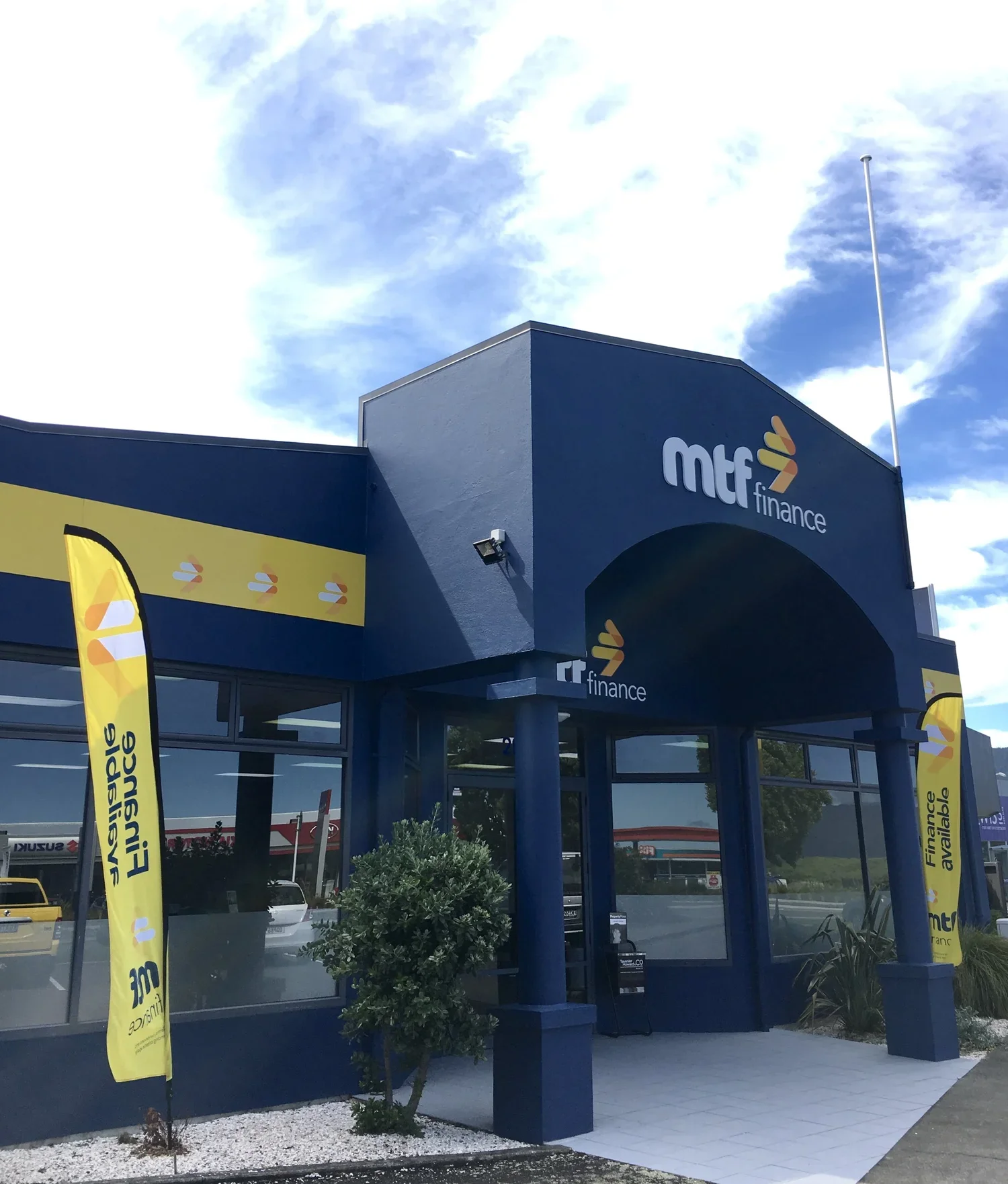 Exterior view of a blue building housing a finance business, with yellow flags displaying the company logo, a small tree in front, and a parking lot visible through the glass windows.
