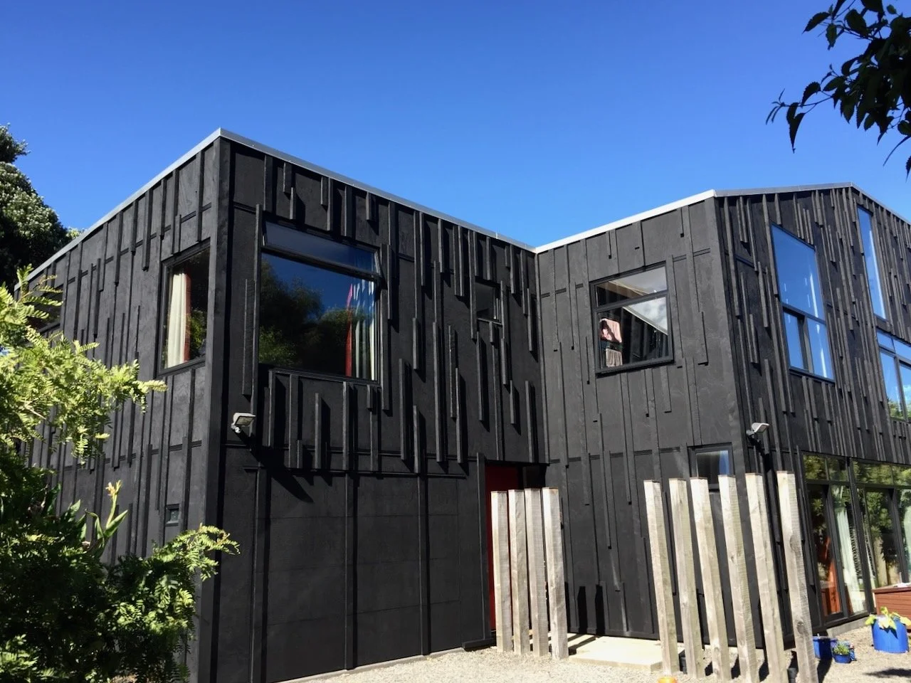 Modern black building with large windows and vertical wood paneling, set against a clear blue sky, with some greenery in the foreground.