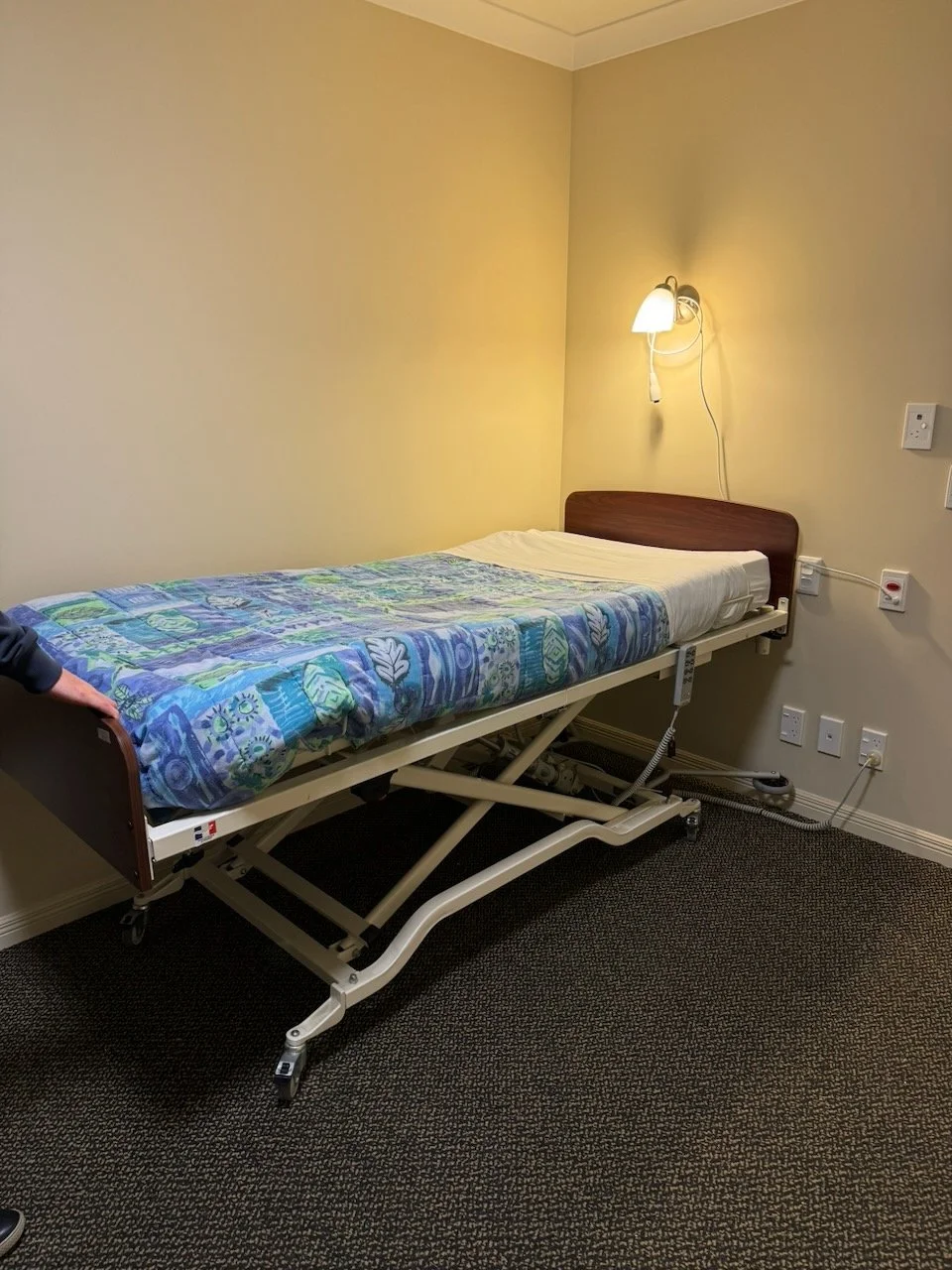 A hospital room with a bed that has a floral cover, placed against a beige wall. There is a wall-mounted lamp above the bed, and multiple electrical outlets on the wall near the bed.
