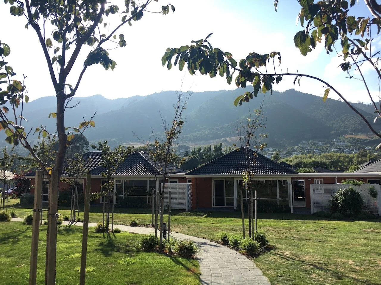 Residential neighborhood with single-story houses, a paved walkway, trees, and hills in the background under a partly cloudy sky.