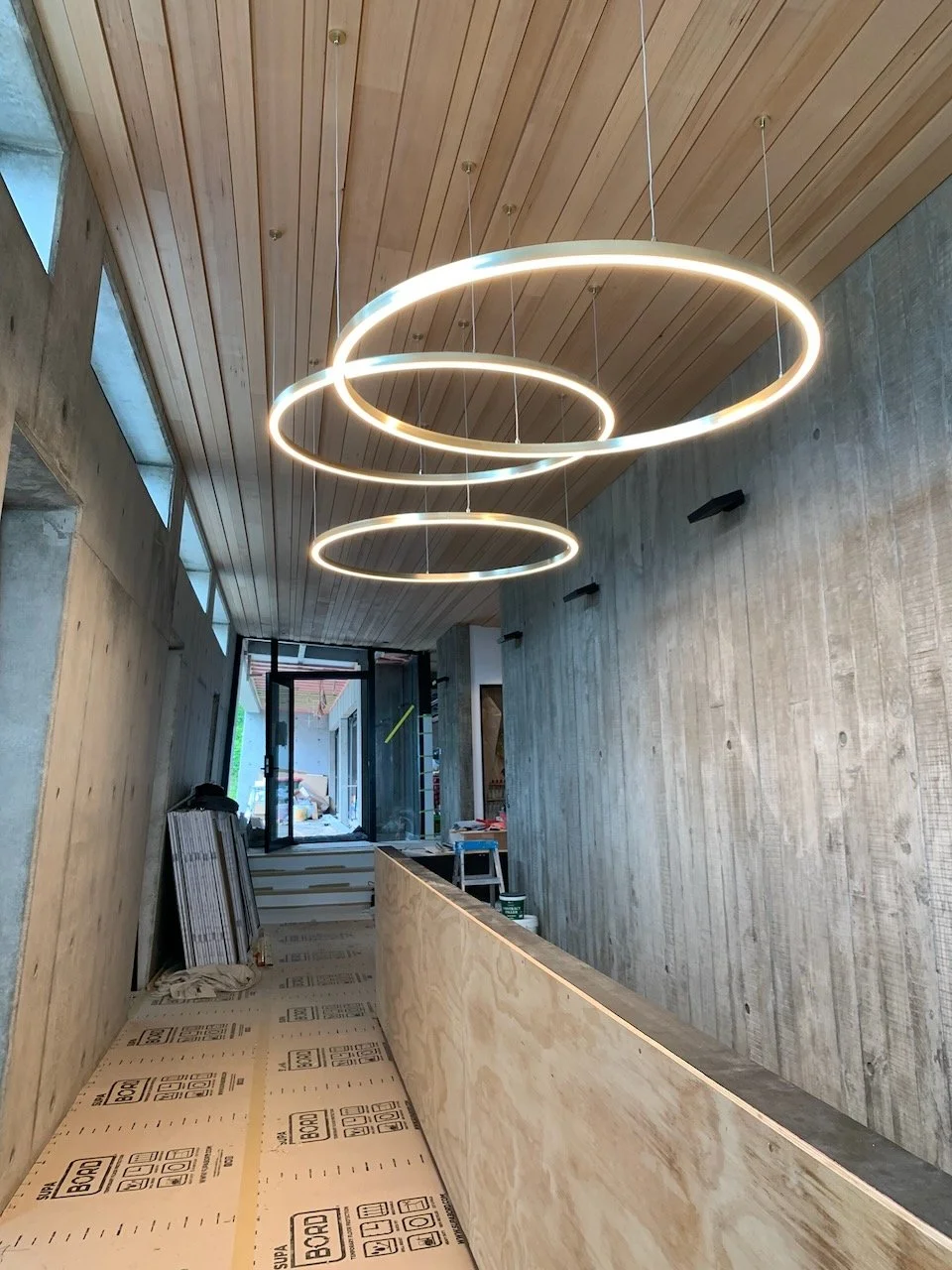 Interior of a building under construction, with a wooden ceiling, concrete walls, modern circular LED light fixtures, and construction materials on the floor.