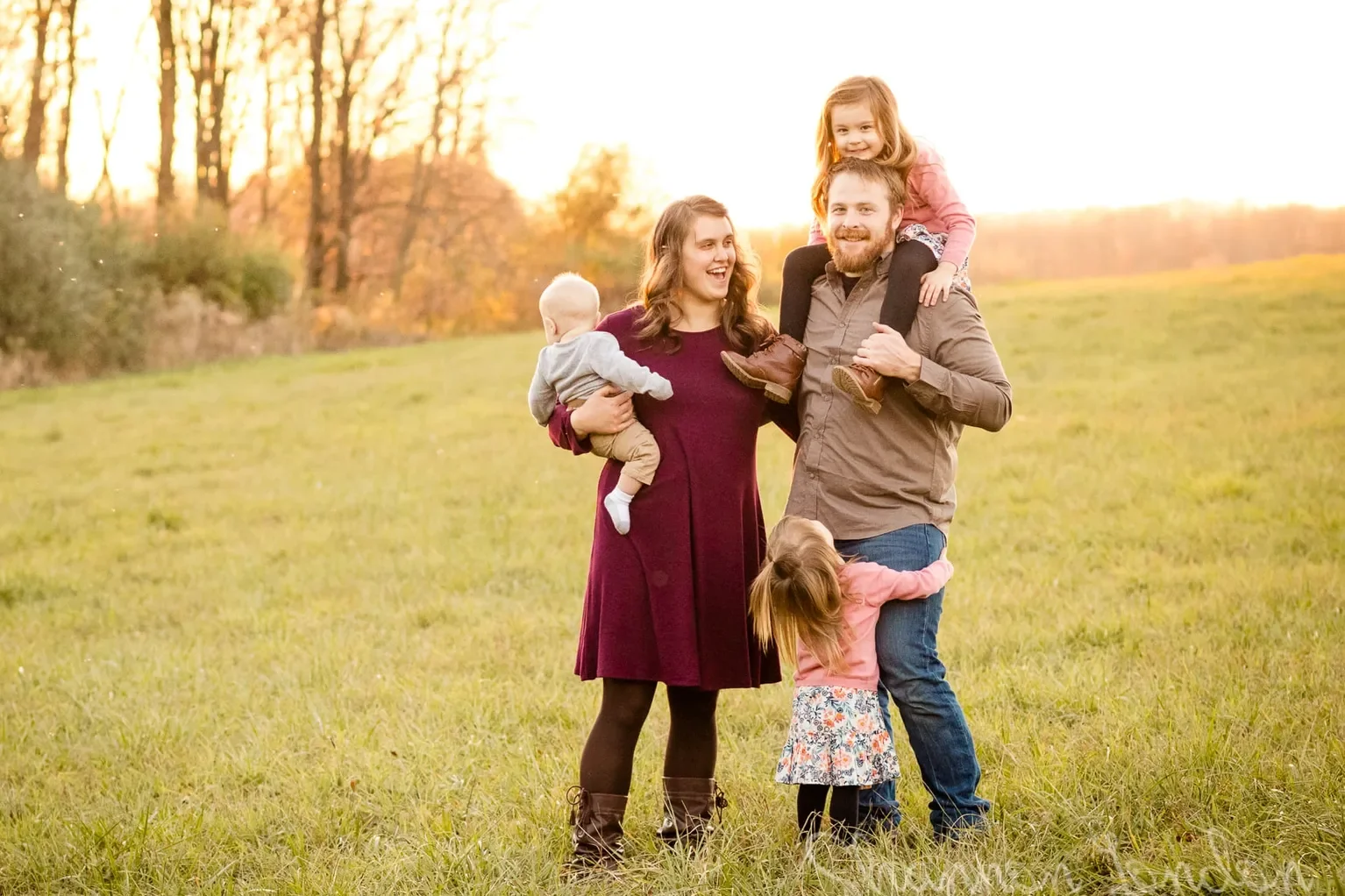 Family of five in a grassy field during sunset. The mother, wearing a maroon dress, holds a toddler. The father, in a brown shirt and jeans, carries a young girl on his shoulders. A little girl hugging his leg is also present, all smiling and enjoying the outdoors.