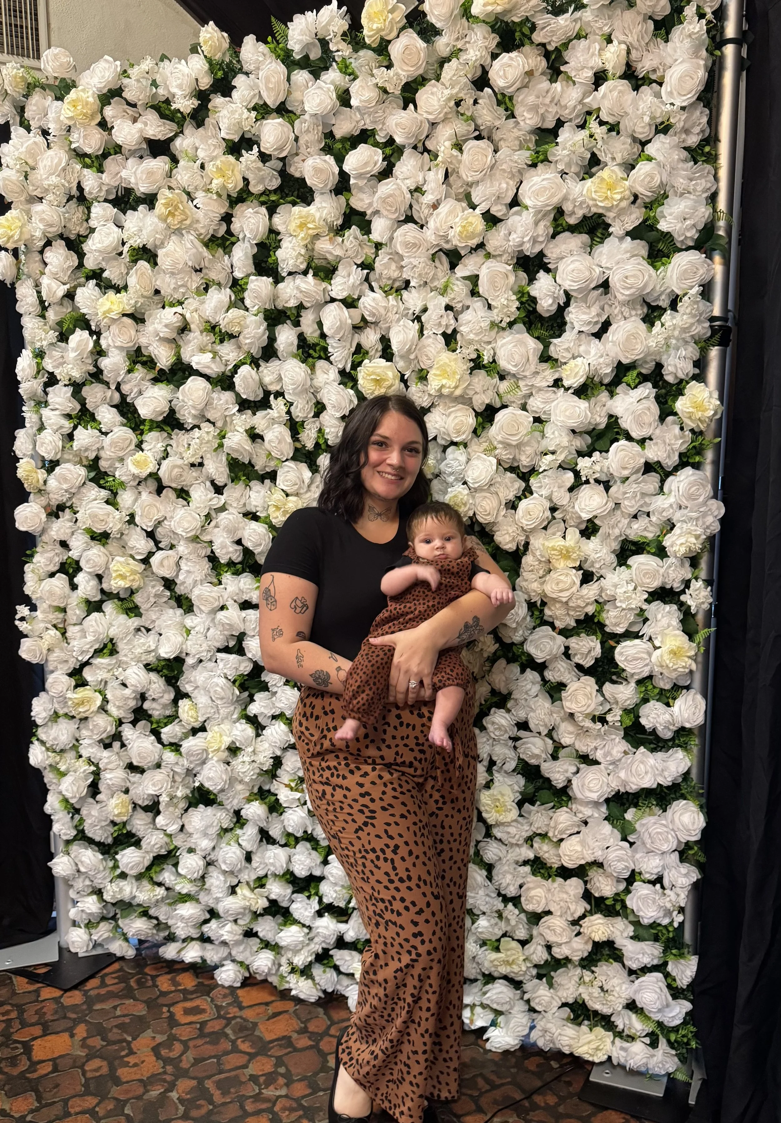 A woman holding a baby, both standing in front of a large floral backdrop made of white roses. The woman has dark hair, tattoos on her arms, and is wearing a black top and leopard print pants. The baby is wearing a brown dress with black spots.