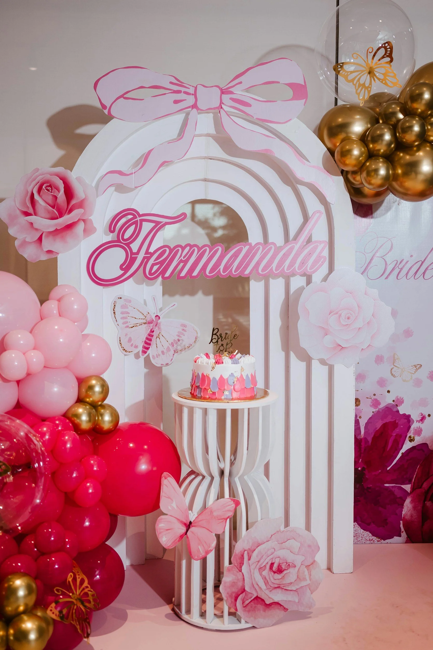 Decorative birthday or celebration display with pink, gold, and white balloons, paper flowers, butterflies, and a cake, featuring a sign with the name 'Fernanda' in pink cursive letters.