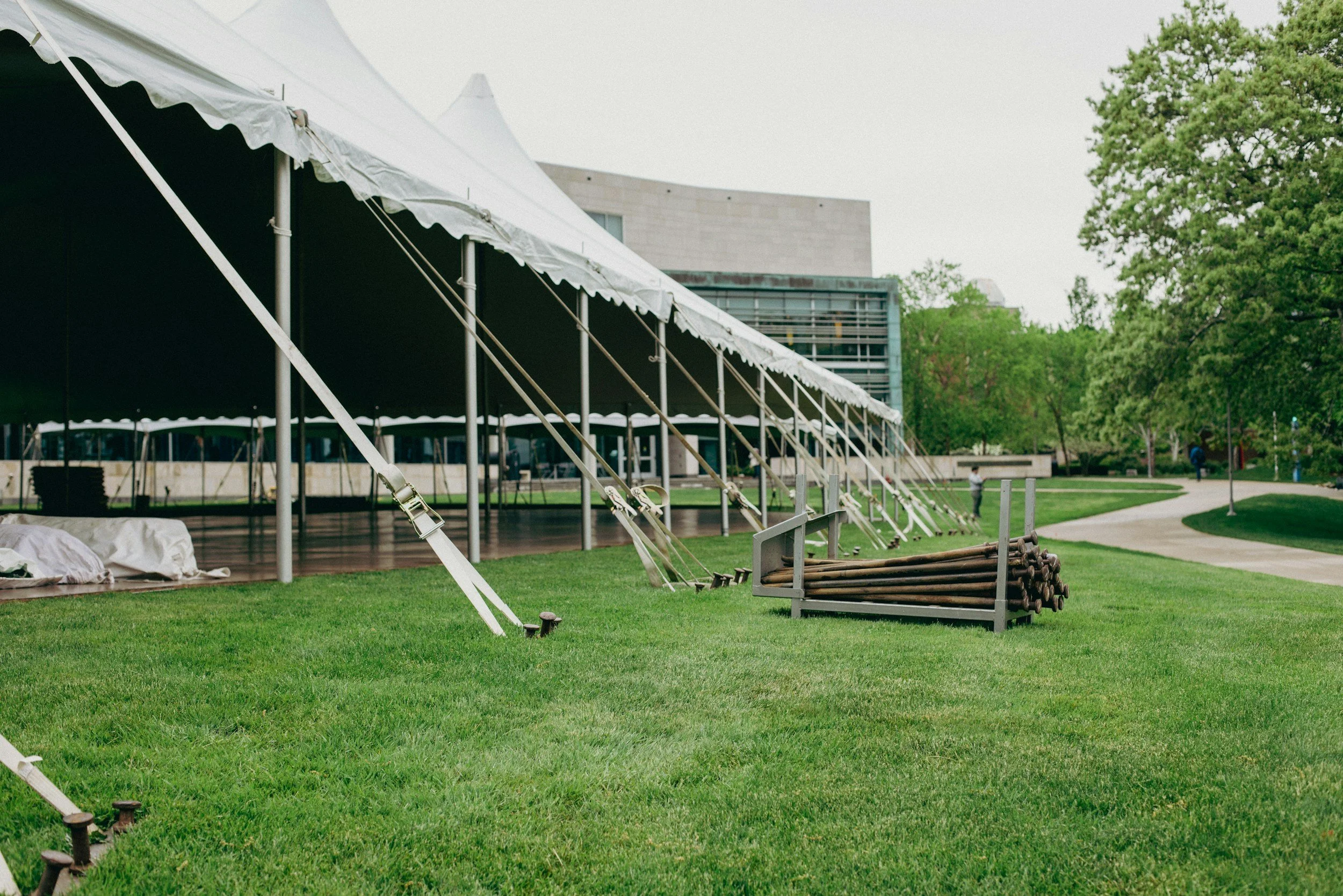 Empty outdoor event tent with ties and poles, wooden benches, and a grassy area with trees and a modern building in the background.