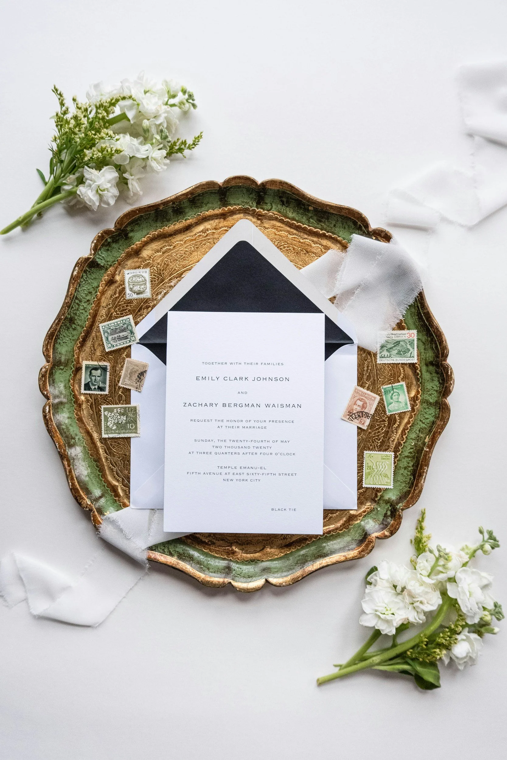 Wedding invitation on a decorative gold and green tray with vintage stamps, surrounded by white flowers on white fabric.