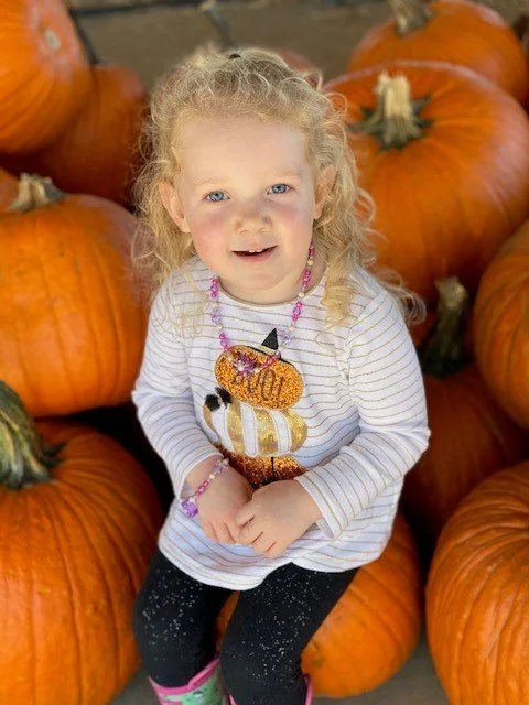 A young girl with blonde curly hair smiling, sitting among large pumpkins at pumpkin patch.