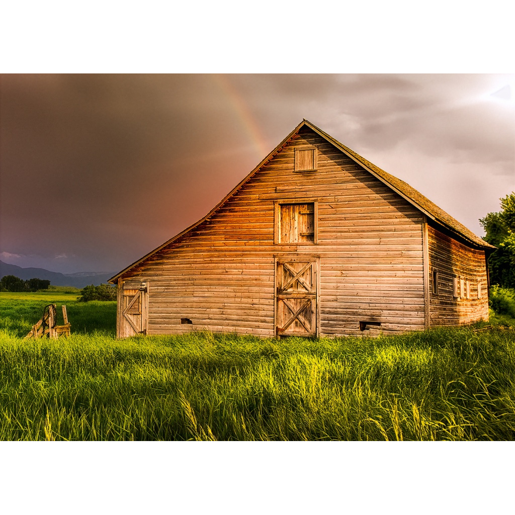 Weathered wooden barn in a green field beneath a dramatic sky with a faint rainbow near Bozeman, Montana, photographed by Tom Lance.