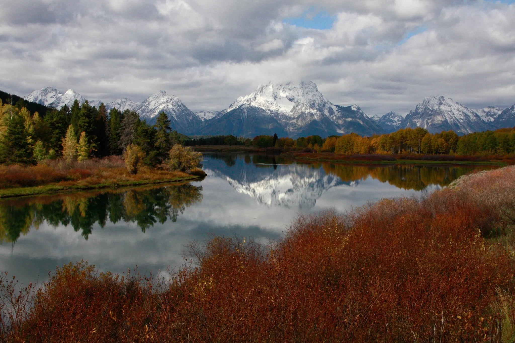 Snow-covered mountains in the background with a cloudy sky, a lake reflecting the mountains and sky, surrounded by autumn trees and bushes in the foreground.