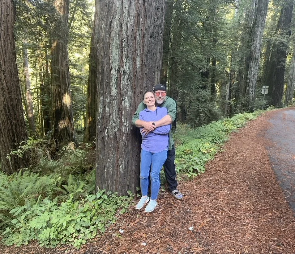 A man and woman smiling and hugging next to a large tree in a forest with tall trees and green foliage, on a paved trail.