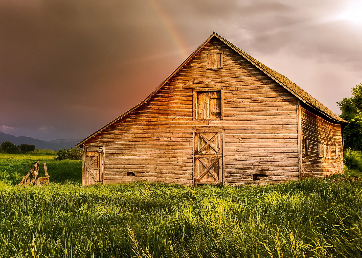 Weathered wooden barn in a green field beneath a dramatic sky with a faint rainbow near Bozeman, Montana.